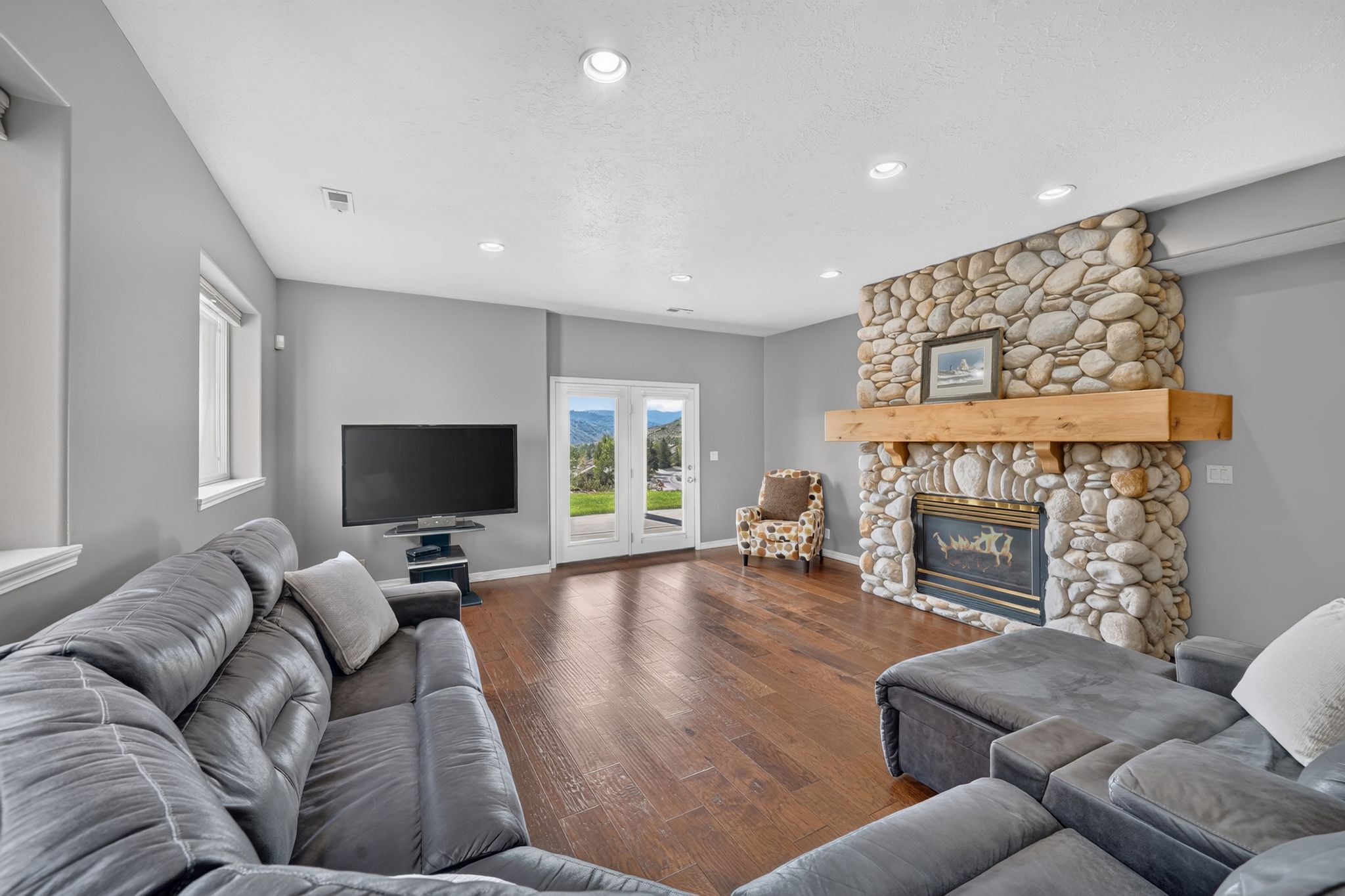 Living area featuring wood-finish flooring, a stone-clad fireplace with a substantial wood mantel, recessed lighting, and double glass doors leading outdoors
