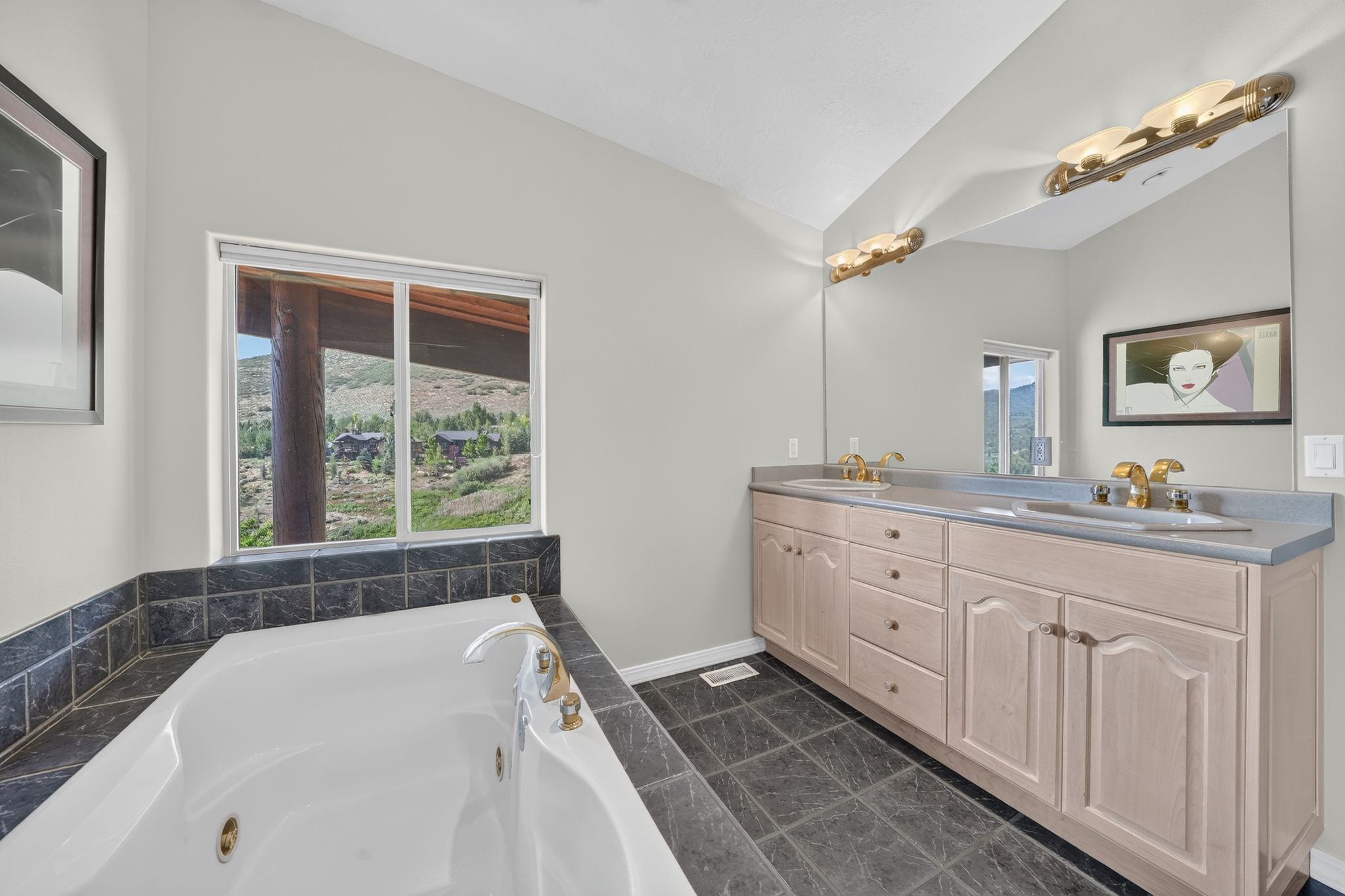 Soaking tub with dark tile surround, dual vanity with light wood cabinetry, gold-tone fixtures, and a large wall-mounted mirror