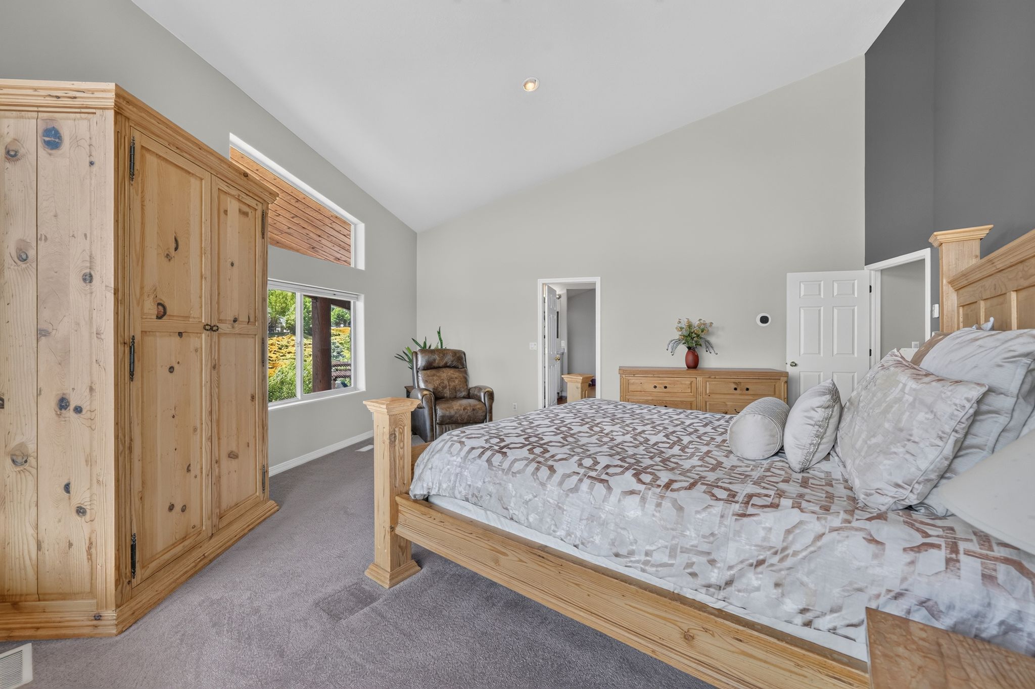 Vaulted ceiling bedroom featuring light gray walls, plush gray carpeting, and a side window with exterior foliage