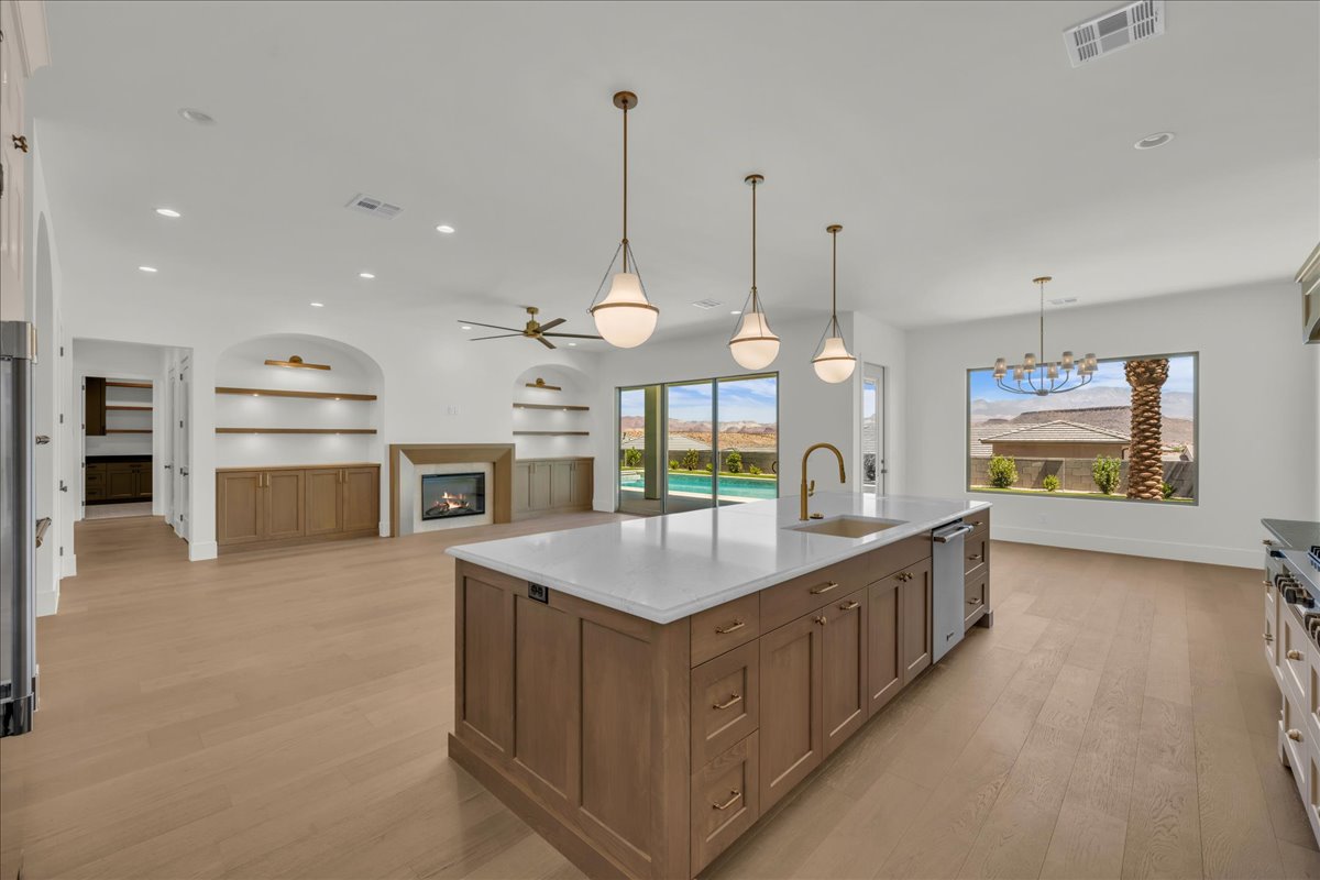 Open-concept living space featuring a substantial kitchen island with a white countertop and undermount sink, light wood-finish flooring, a contemporary fireplace, built-in shelving with lower cabinetry, and large windows overlooking a pool and desert lan