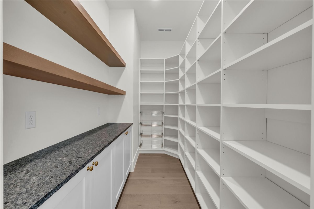 Walk-in pantry featuring extensive white shelving, wood-finish floating shelves, a dark speckled countertop, white cabinetry with gold-tone hardware, and wood-finish flooring