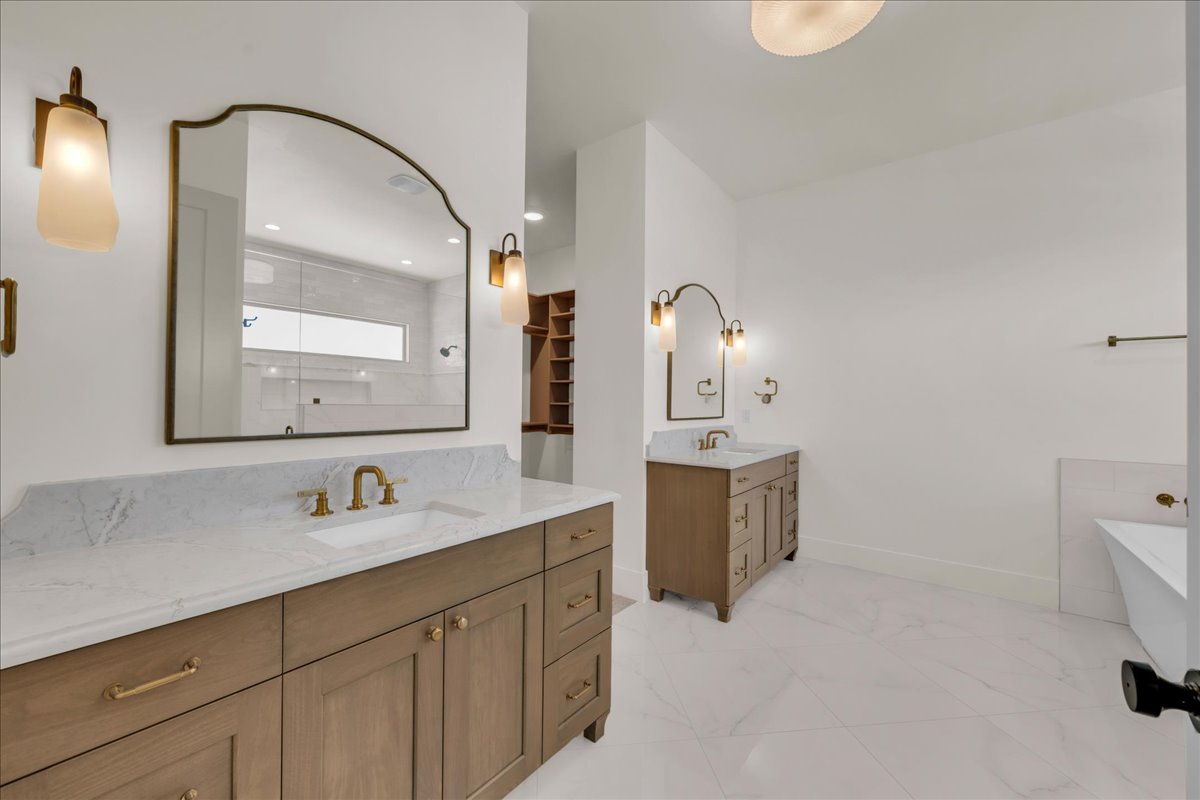 Spacious bathroom featuring dual wood-finish vanity cabinets with stone countertops, a freestanding tub, and large format floor tiling