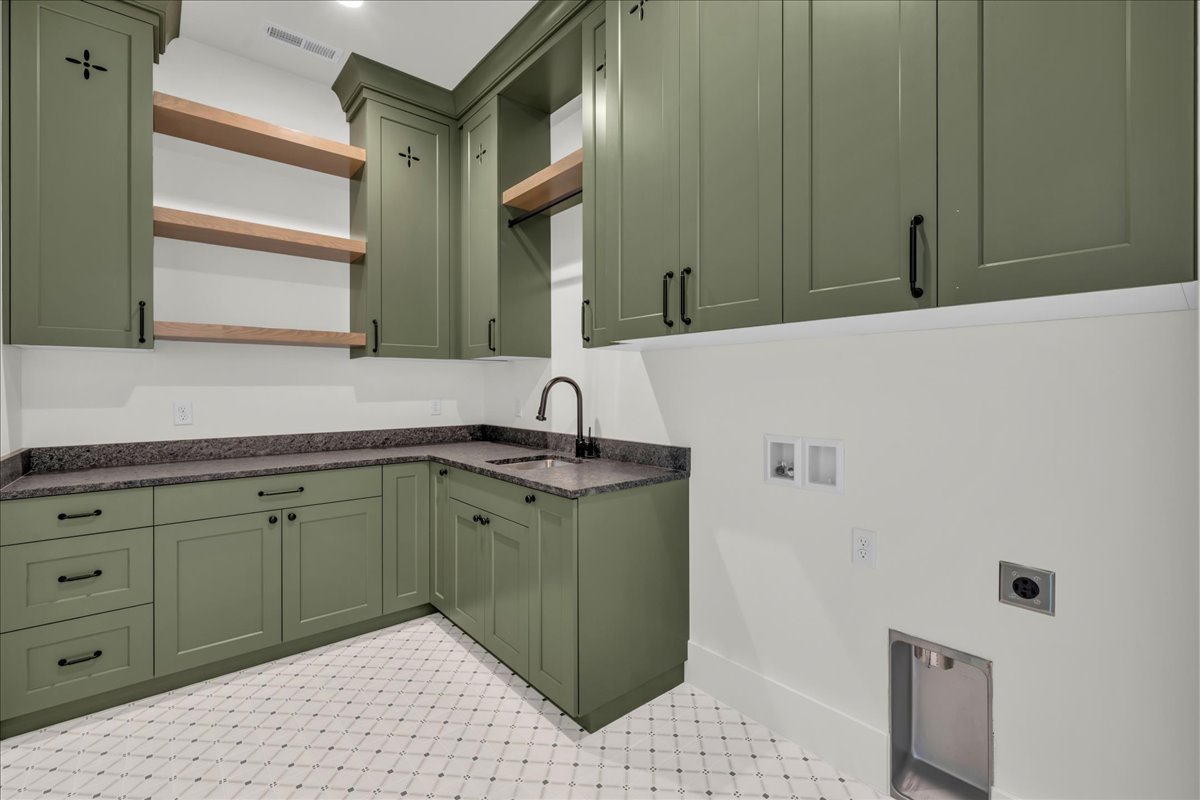 Laundry area featuring olive green cabinetry, dark stone countertops, a corner sink with a black faucet, and white patterned floor tiling