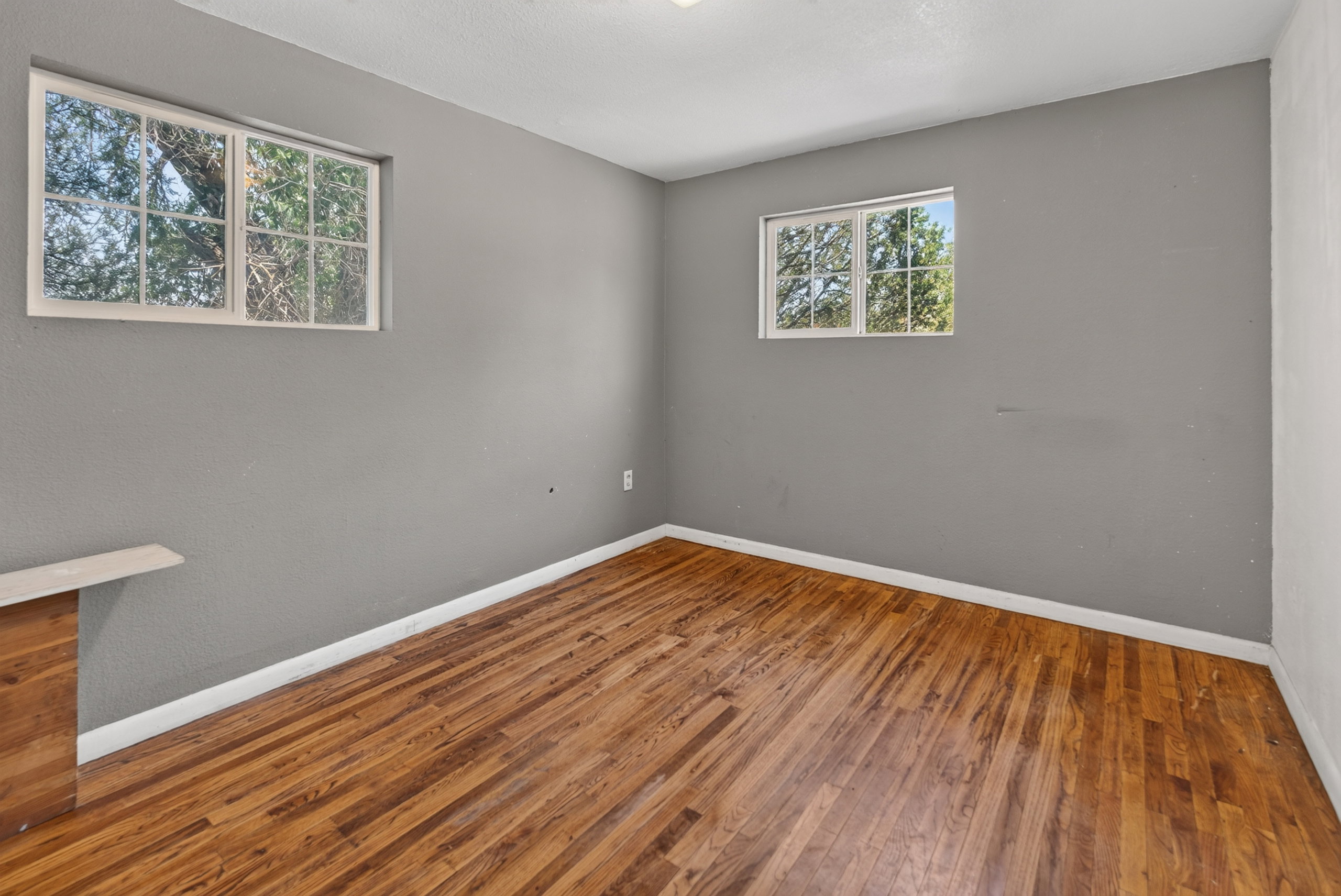 Room featuring two windows, wood-finish flooring, light gray wall paint, white baseboards, and a built-in wood-finish cabinet with a light-toned top