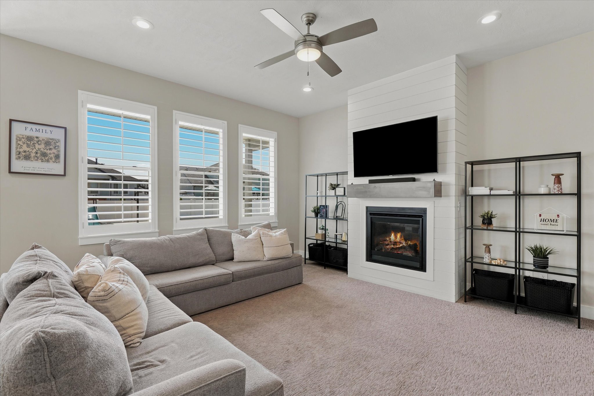 Spacious living area featuring a fireplace with a shiplap accent wall, built-in shelving, a ceiling fan, recessed lighting, and triple windows with plantation shutters