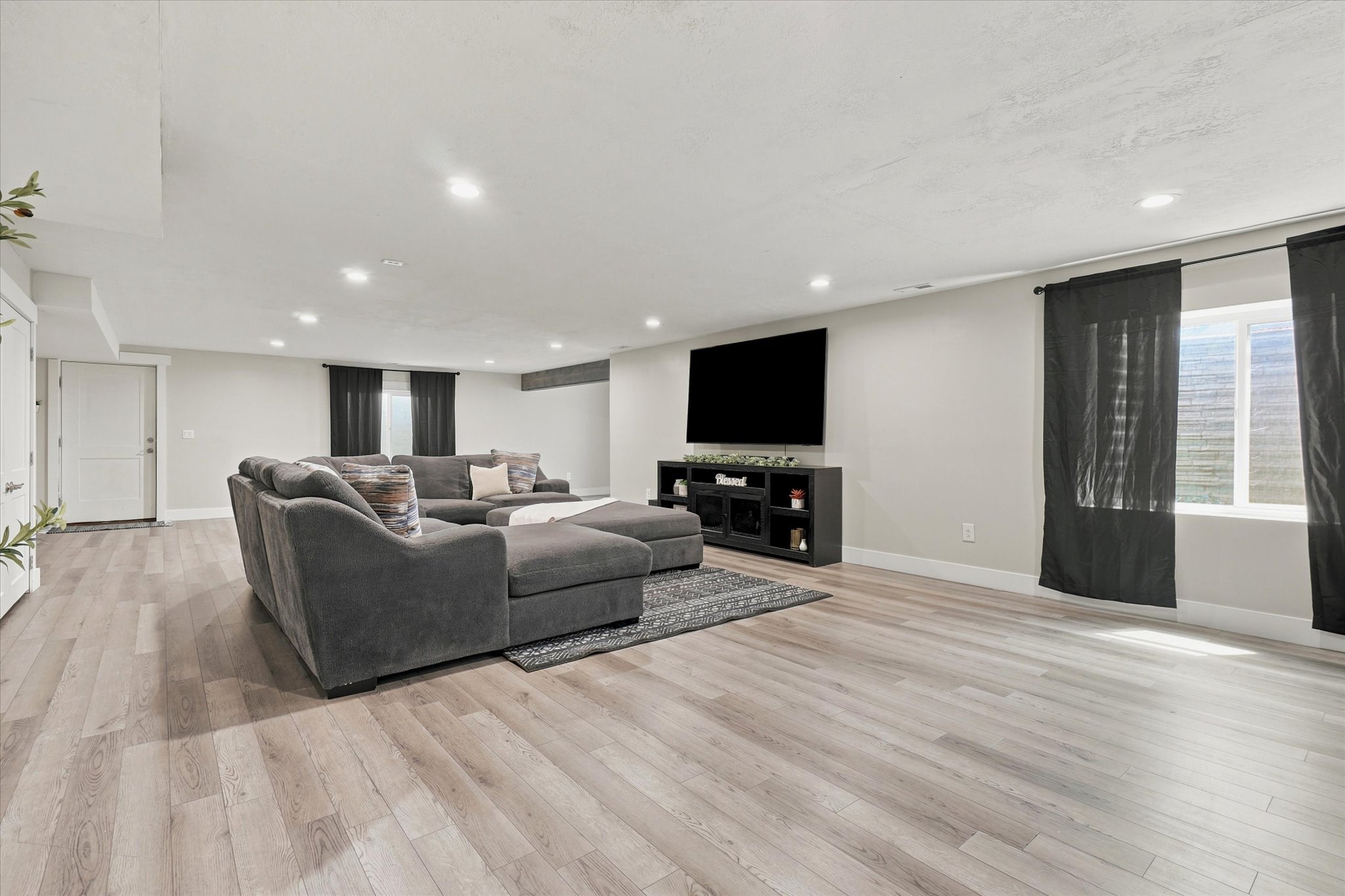 Spacious living area featuring light wood-finish flooring, recessed lighting, white walls, and a large window