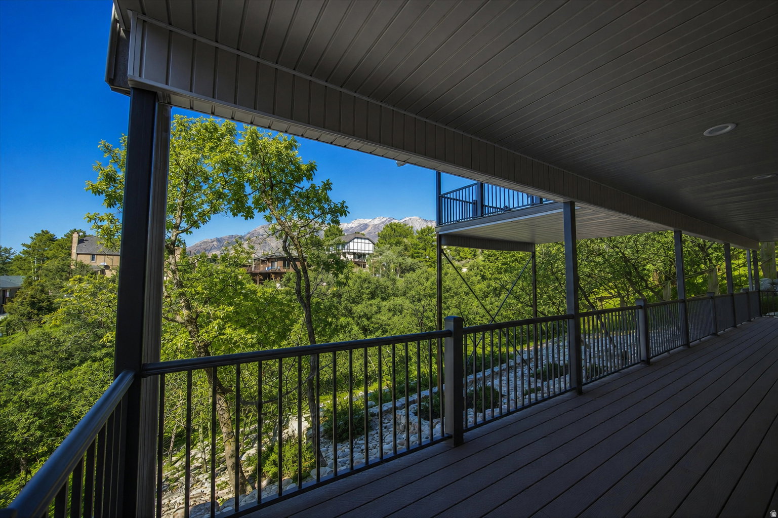 Expansive outdoor deck featuring dark composite decking, black metal railings, a soffit ceiling with recessed lighting, and a partial mountain backdrop