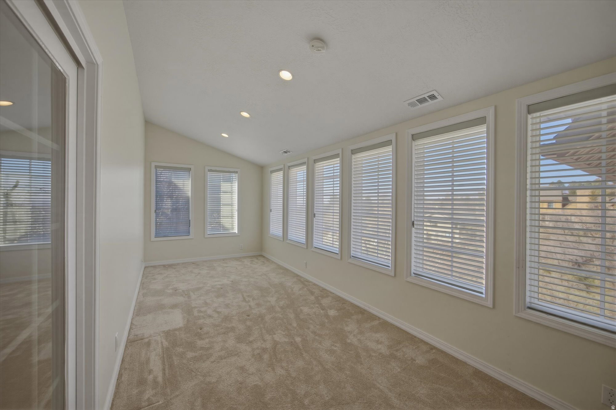 Spacious room featuring multiple windows with blinds, recessed lighting, a vaulted ceiling, light beige walls, and neutral-toned carpeting