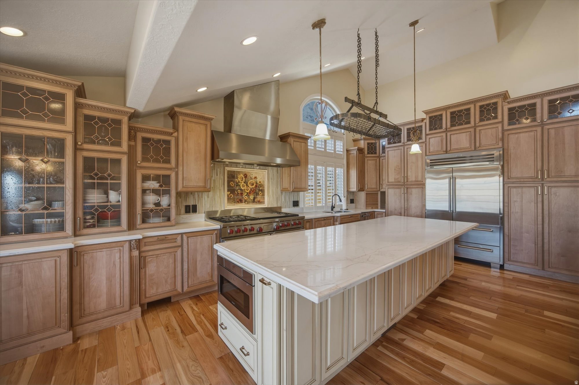Gourmet kitchen featuring a large central island with integrated microwave, stainless steel appliances, extensive cabinetry with glass-front accents, wood-finish flooring, and vaulted ceilings