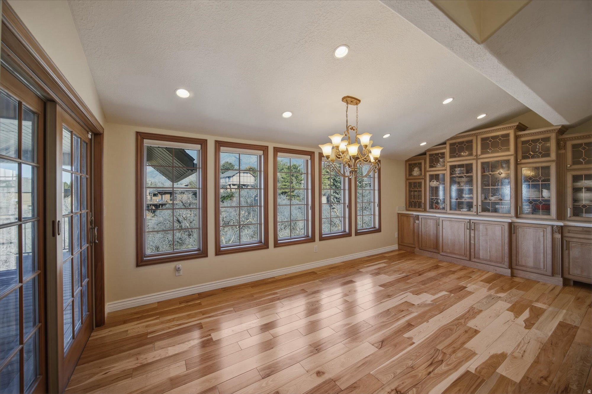 Interior room featuring wood-finish flooring, a wall of built-in cabinetry with glass-front uppers, multiple large windows with wood trim, recessed lighting, and a decorative chandelier