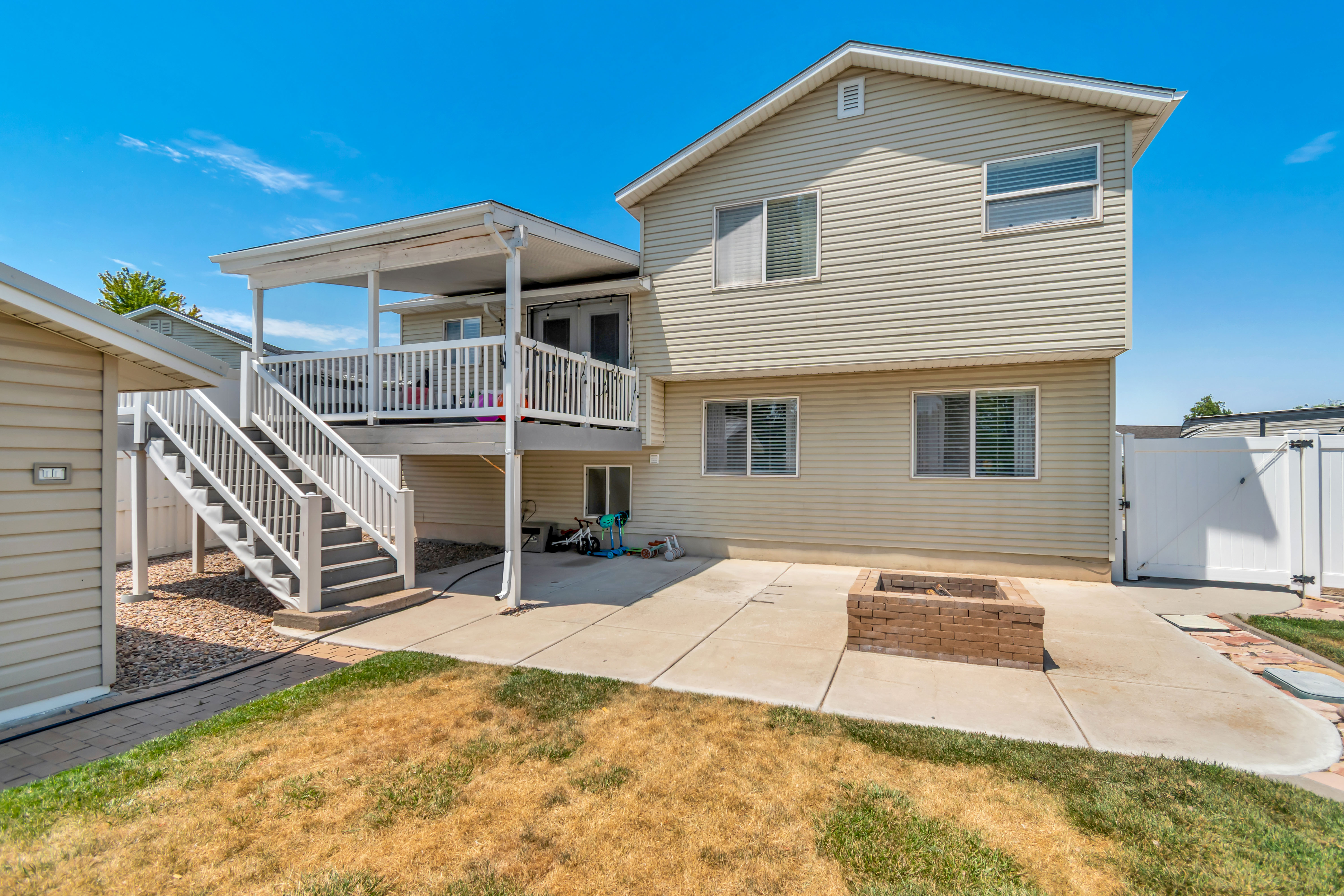 Rear exterior featuring a covered deck with white railings and dual staircases, a concrete patio, and a built-in brick fire pit