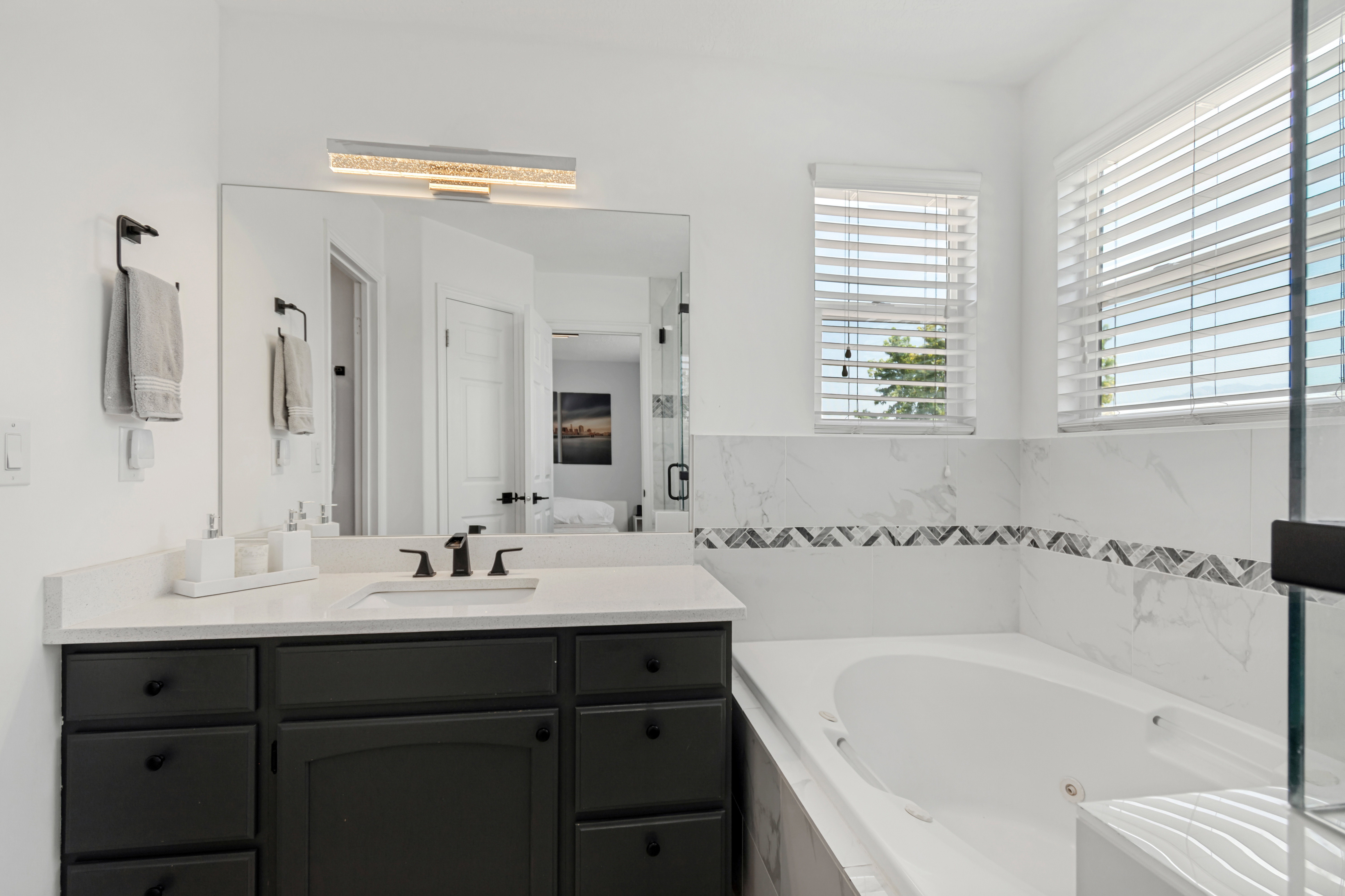 Spacious bathroom featuring a built-in soaking tub, a vanity with a white countertop and dark cabinetry, and a large wall-mounted mirror with an integrated light fixture