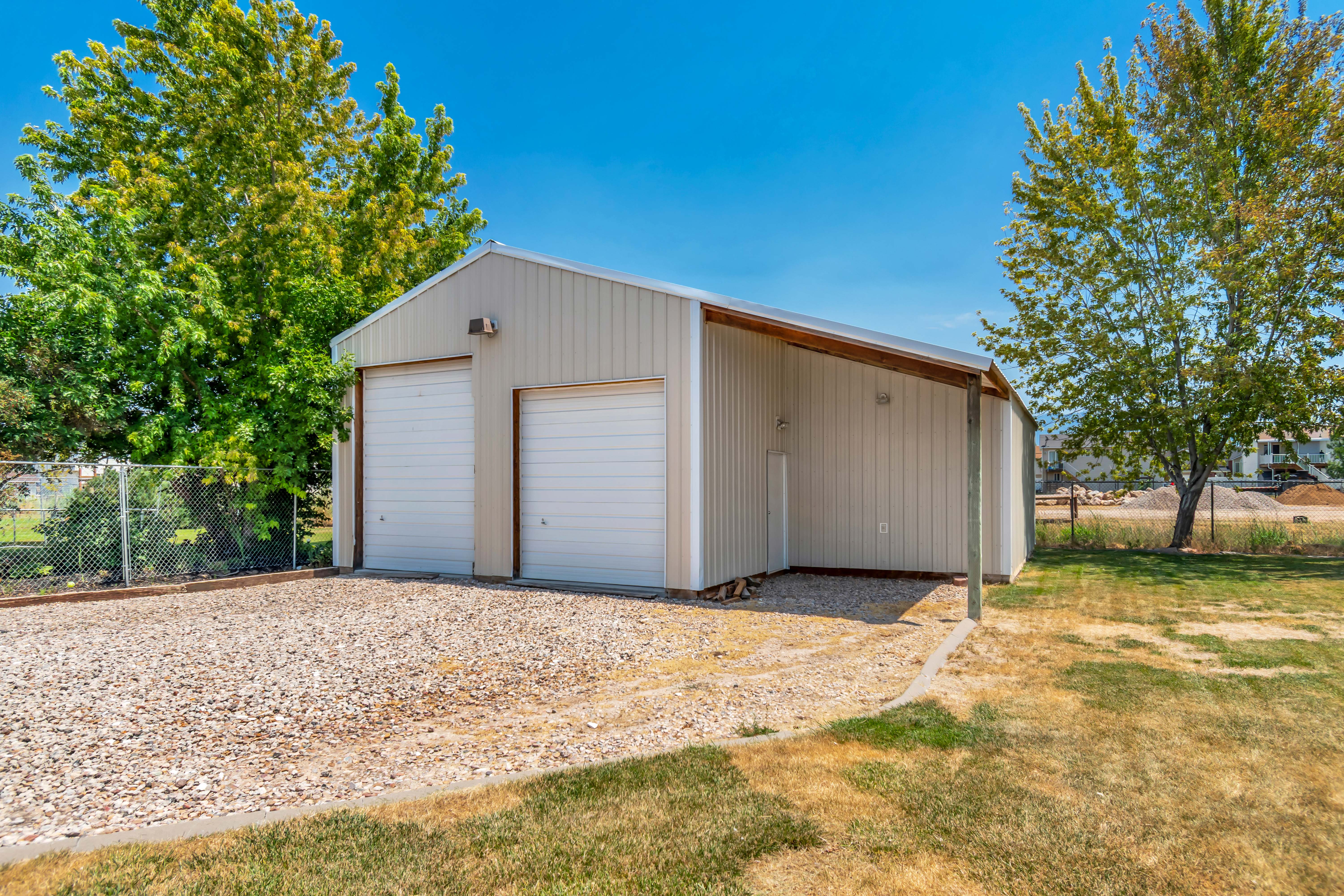 Detached metal garage with two roll-up doors, an extended carport, and a gravel driveway