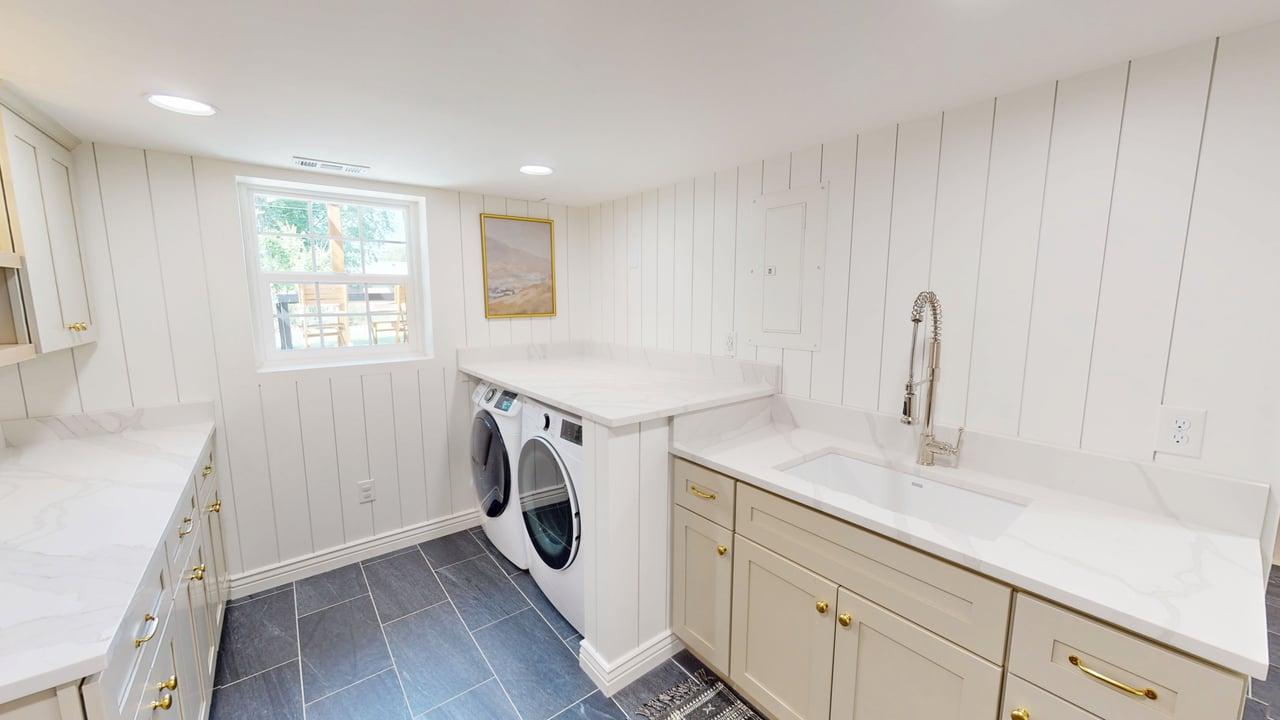 Laundry room featuring white shiplap walls, dark tile flooring, a window, and extensive countertop space with an undermount sink and light-toned cabinetry