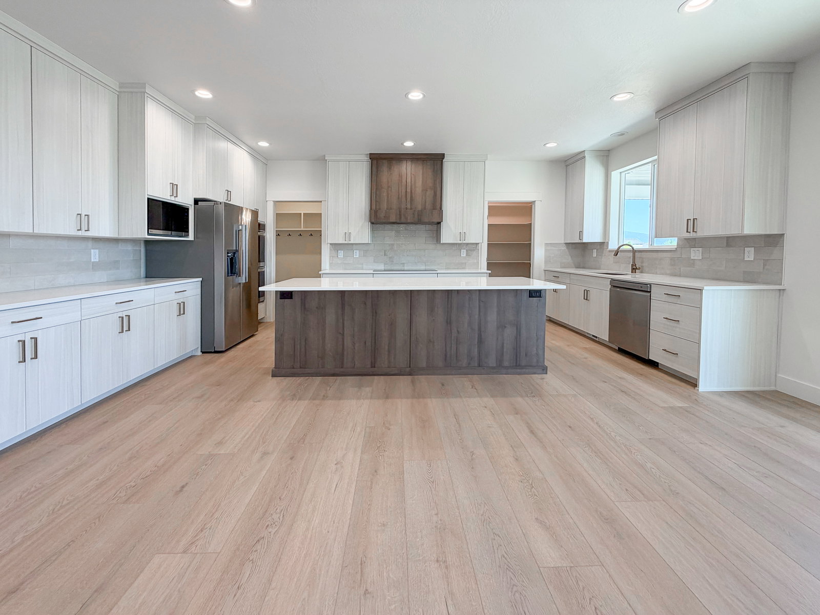 Open-concept kitchen featuring an expansive central island with a wood-finish base, light wood-finish flooring, white perimeter cabinetry with brushed nickel hardware, light countertops, and a subway tile backsplash