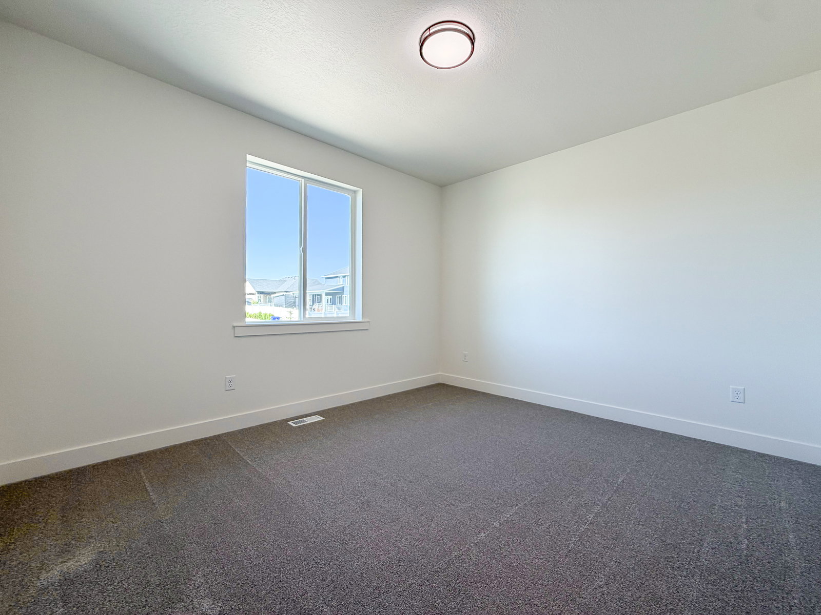 Bright interior space featuring a window with exterior views, light-toned walls, dark gray carpeting, crisp white baseboards, and a flush-mount ceiling light fixture