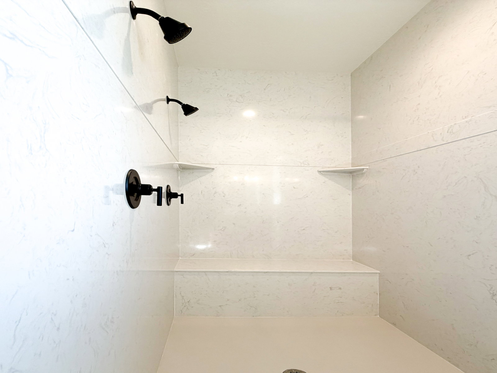 Spacious shower stall featuring a built-in bench, two matte black showerheads, integrated corner shelves, and light-toned wall panels