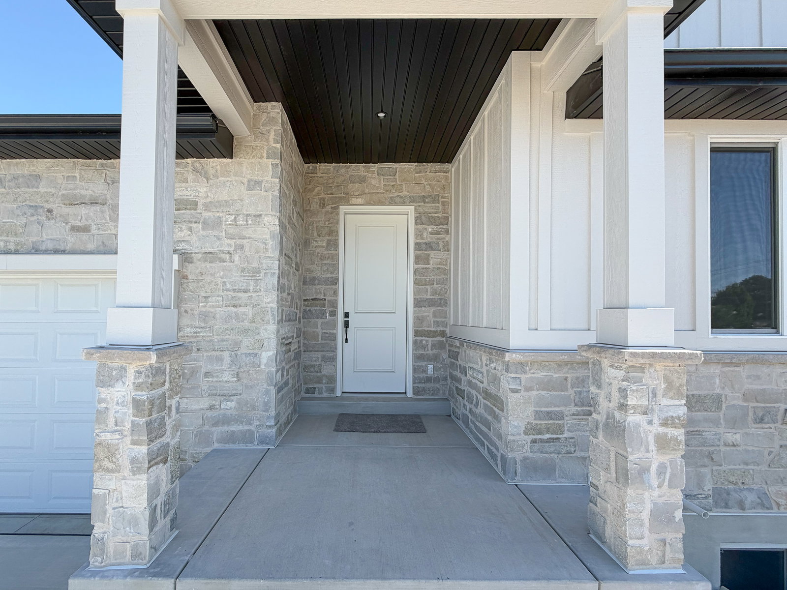 Entryway featuring a white door with black hardware, stone accents, white vertical siding, and a contrasting dark soffit