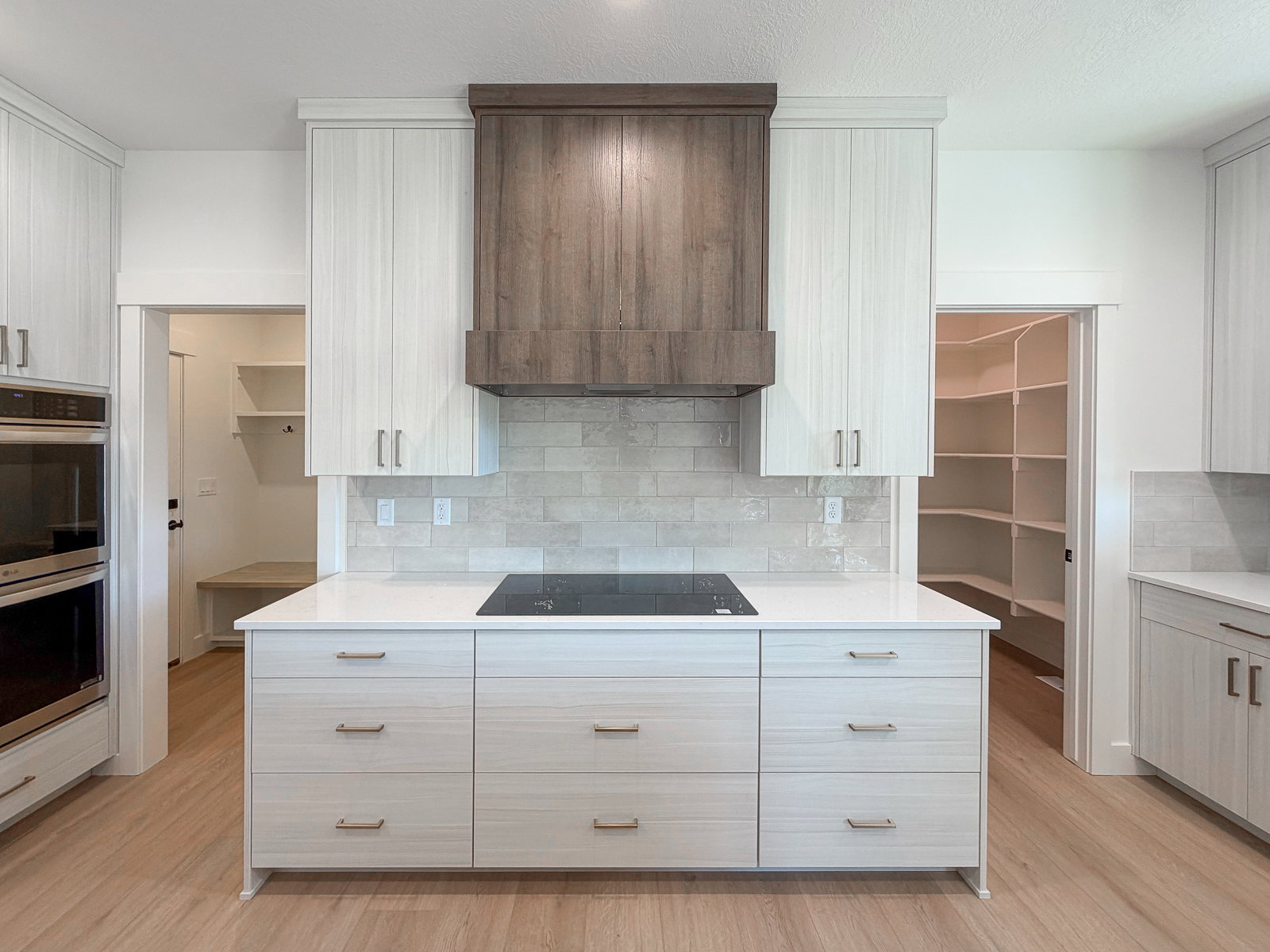 Kitchen featuring a central island with an integrated cooktop, light wood-finish cabinetry, a contrasting wood-finish range hood, double wall ovens, and a light-toned tile backsplash