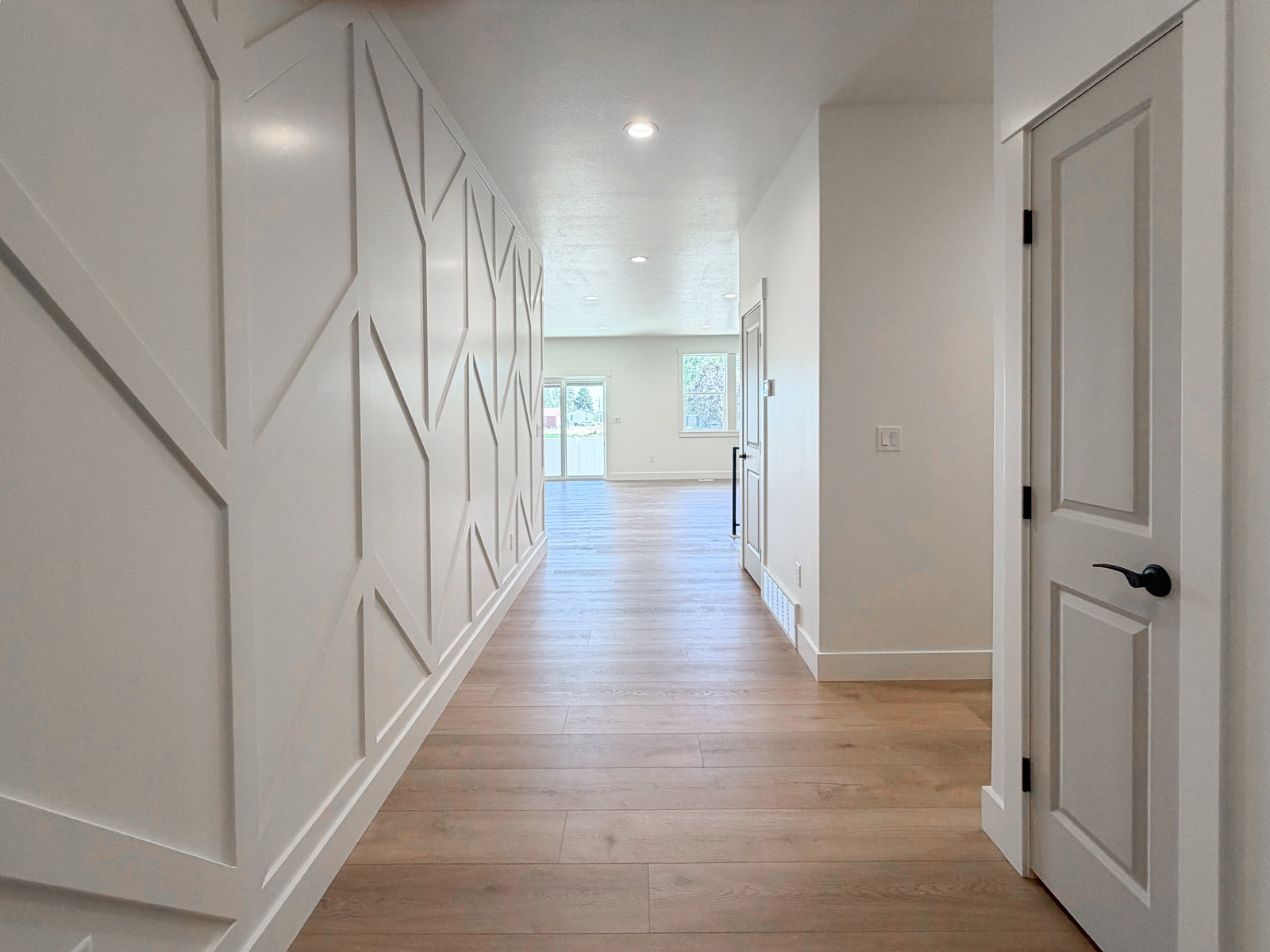 Hallway featuring a decorative accent wall with geometric paneling, wood-finish flooring, white baseboards, recessed lighting, and a paneled interior door with matte black hardware