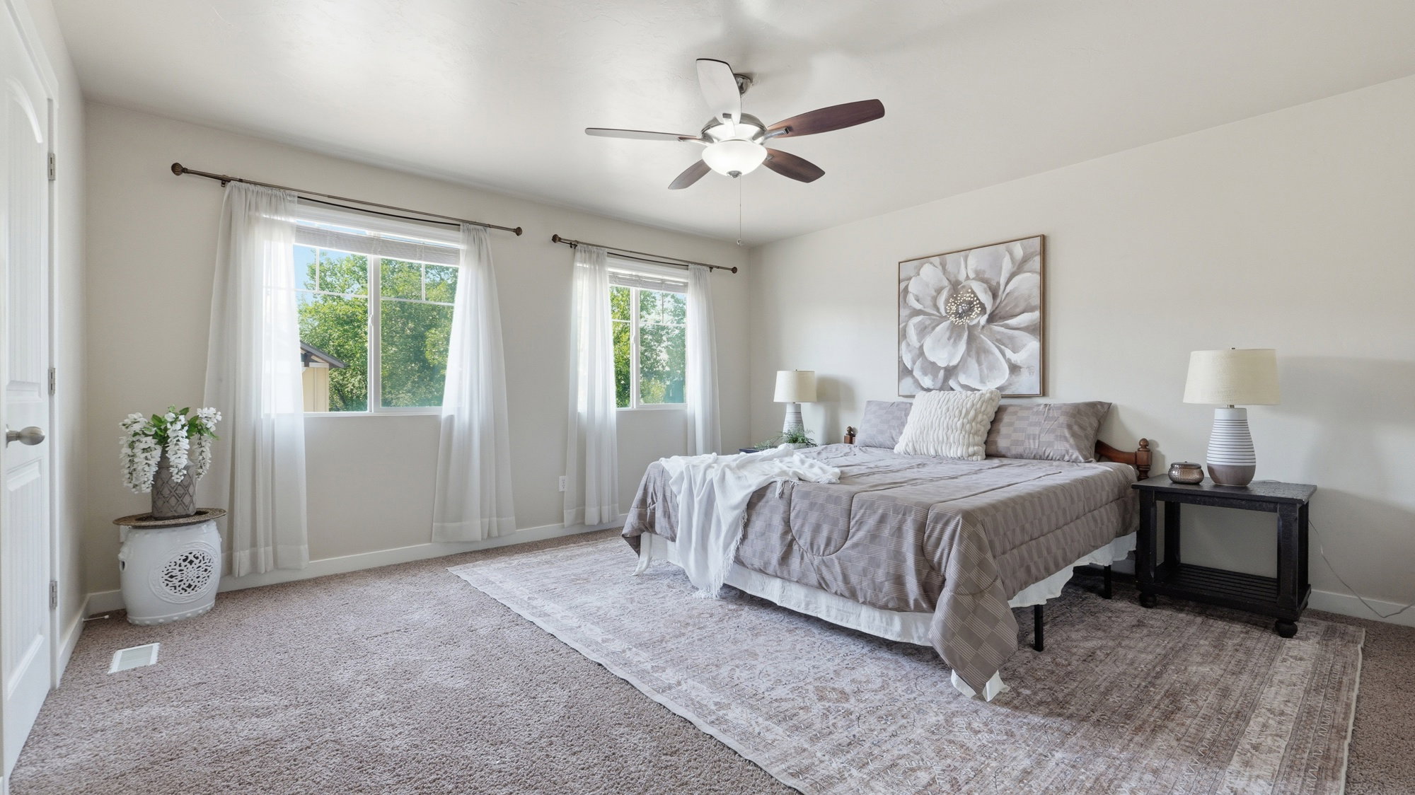Spacious bedroom featuring neutral carpeting, two large windows, a ceiling fan with integrated lighting, and a solid core door