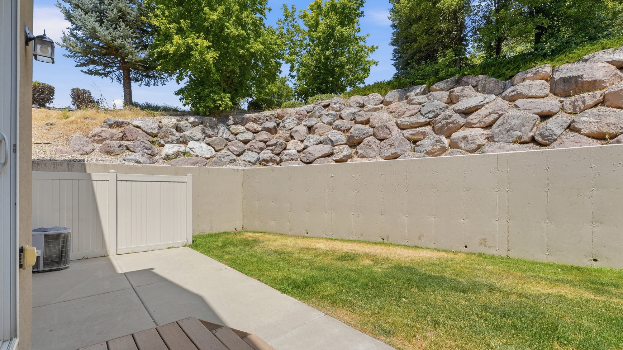 Private patio area featuring a concrete slab, a patch of green lawn, and a stone retaining wall