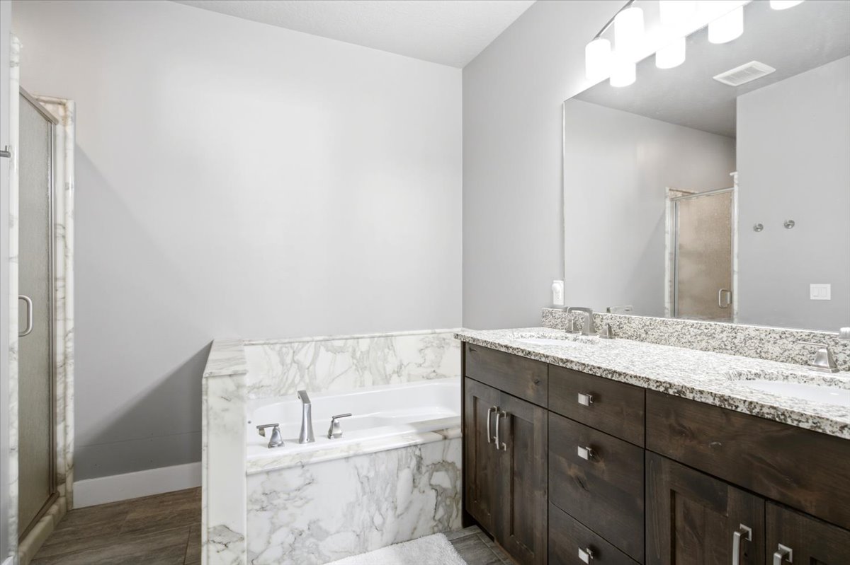 Soaking tub encased in white marble, dual sink vanity with stone countertop, dark wood-finish cabinetry, and a glass-enclosed shower