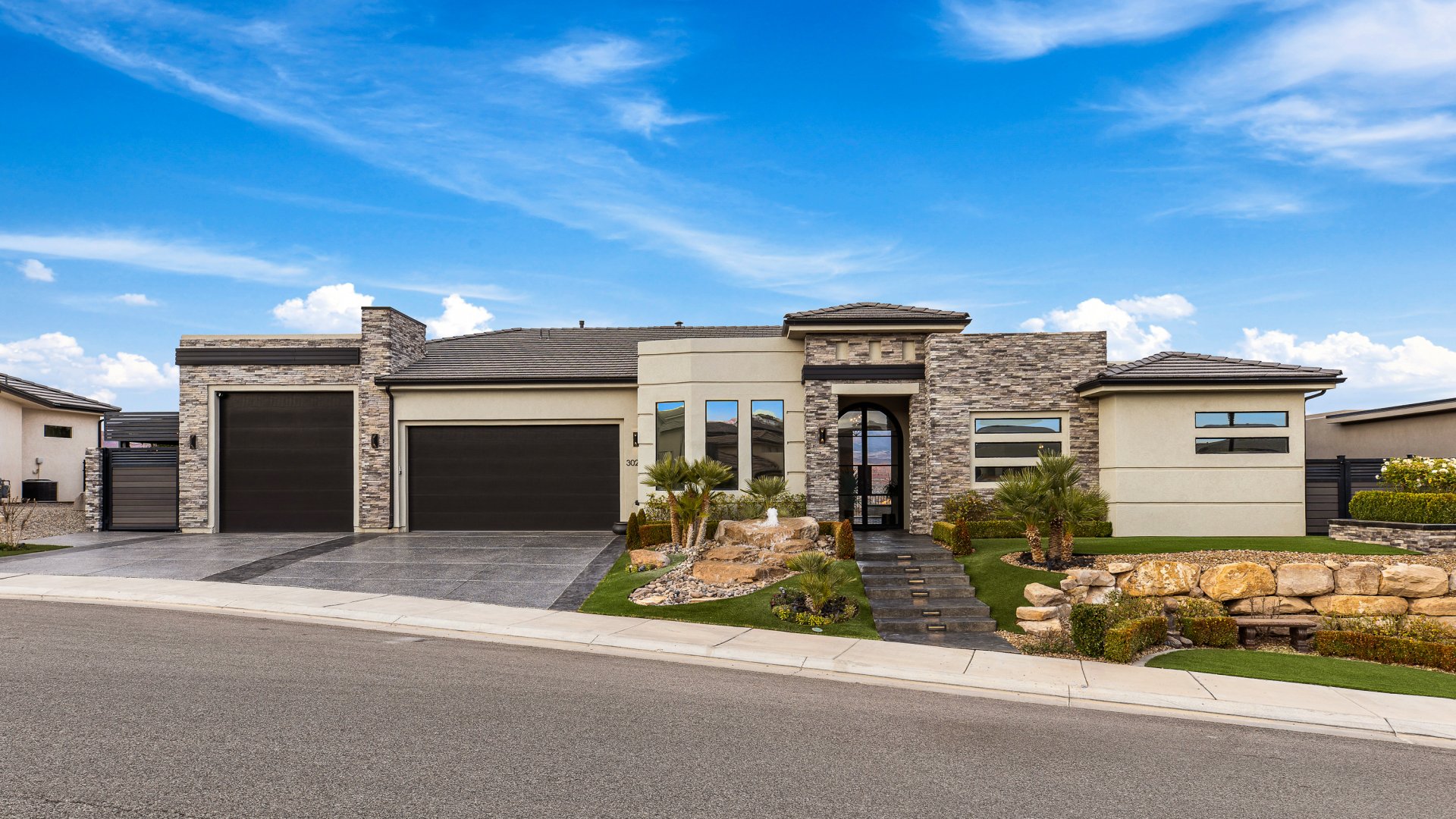 Modern stucco and stone facade featuring a multi-car garage, arched entry door, and tiered landscaping with natural rock elements