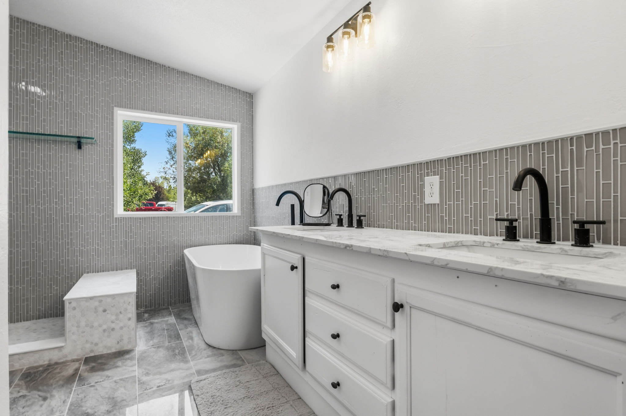 Contemporary bathroom featuring a freestanding tub, a dual vanity with marble-finish countertops, black hardware and fixtures, and a glass tile accent wall