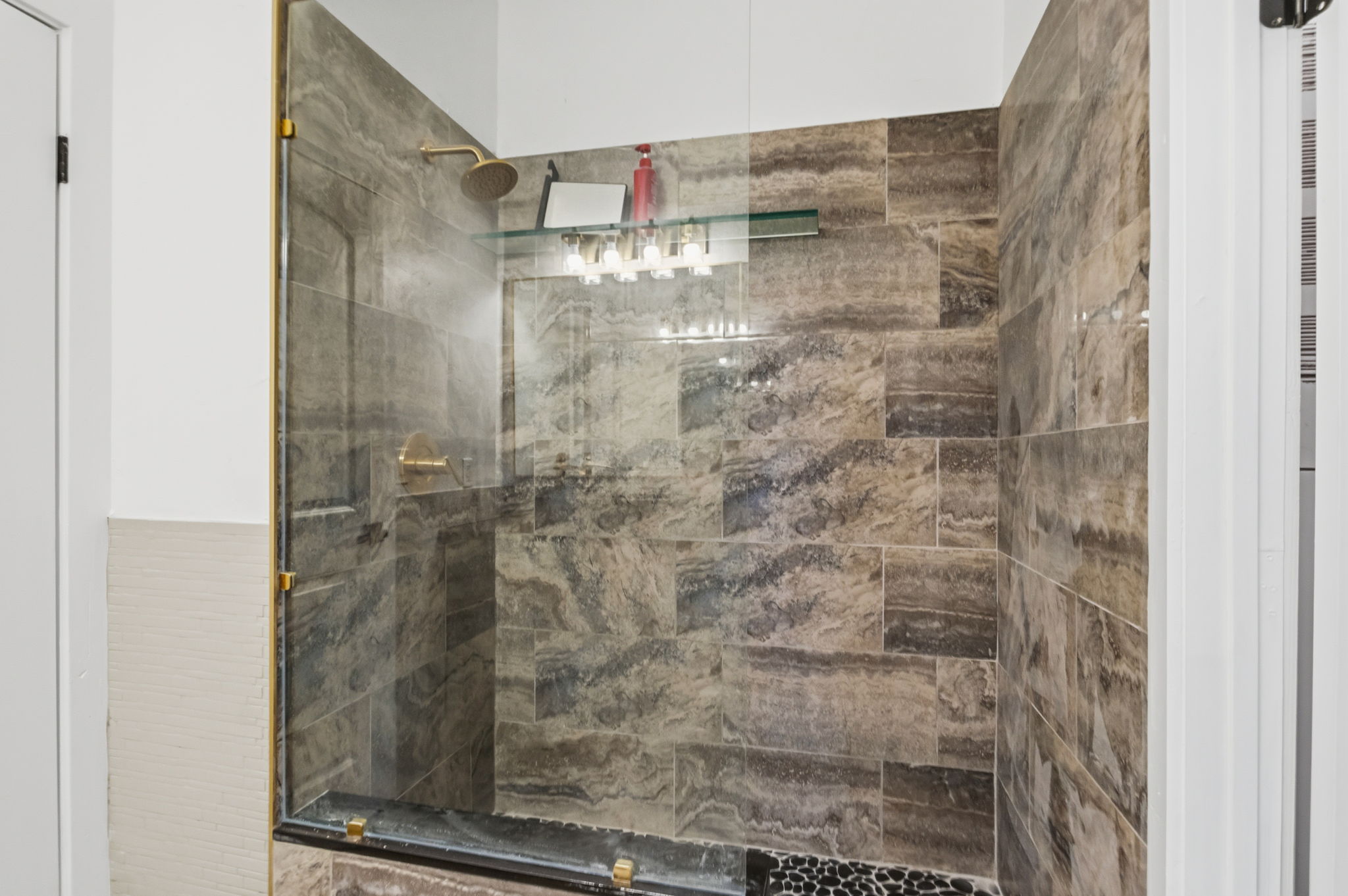 Walk-in shower featuring glass enclosure with brass-finish hardware, large format stone tile walls, a pebble-finish floor, and a built-in glass shelf