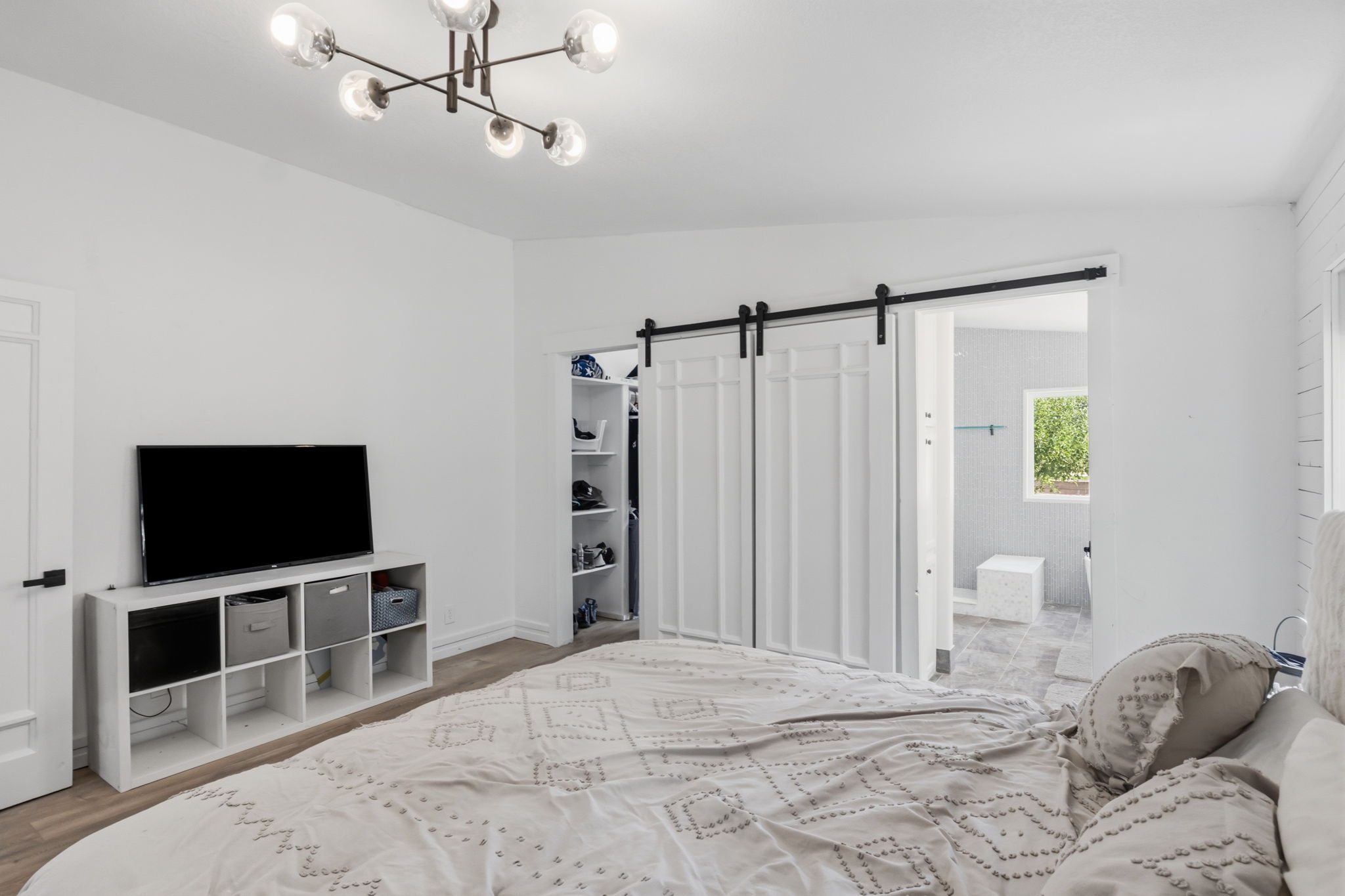 Bedroom featuring modern ceiling light fixture, white walls, wood-finish flooring, and a barn door leading to an adjacent bathroom