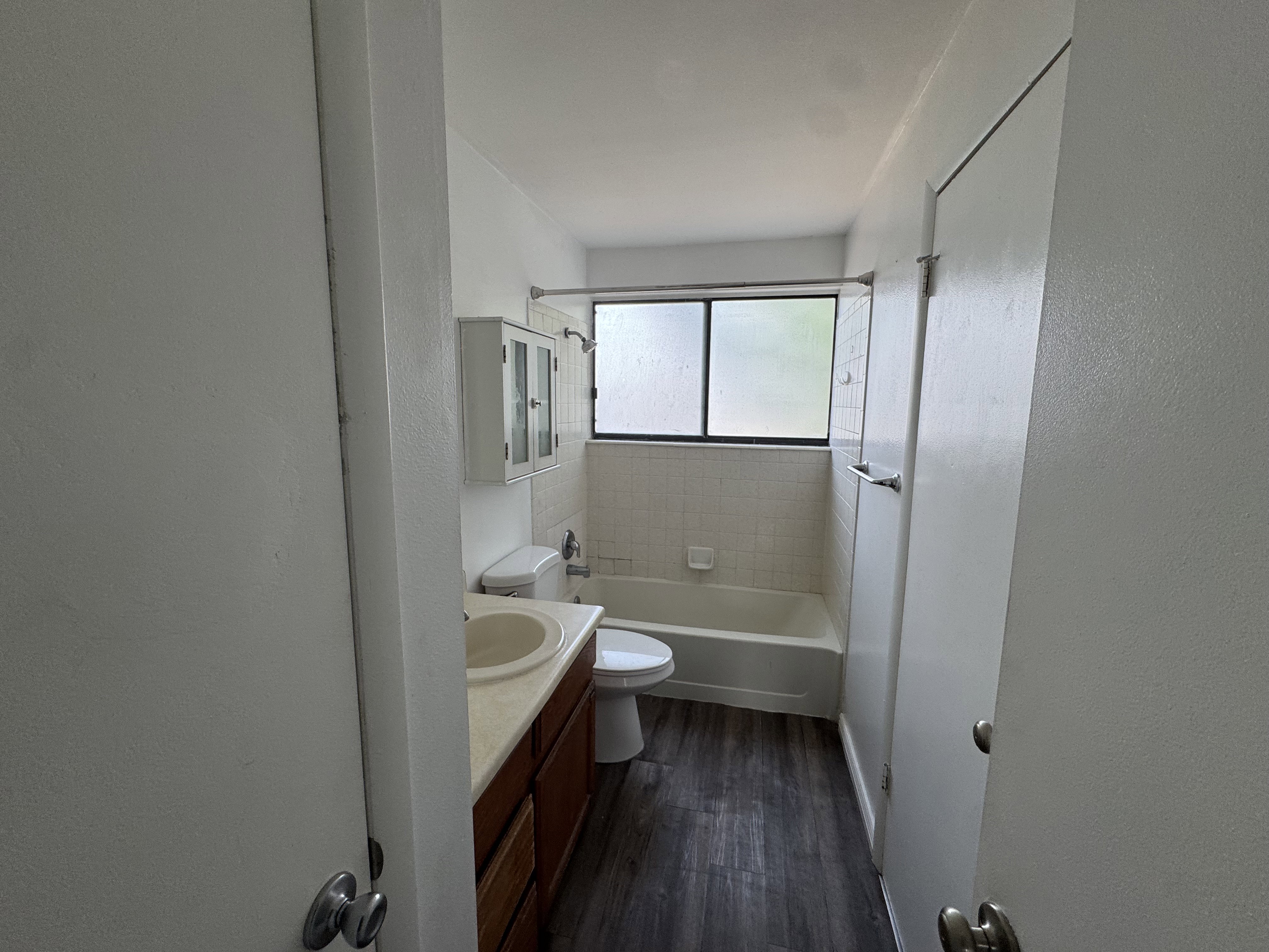 Bathroom featuring wood-finish flooring, a white bathtub with tile surround, a built-in vanity with light-colored countertop and integrated sink, and a window with frosted panes