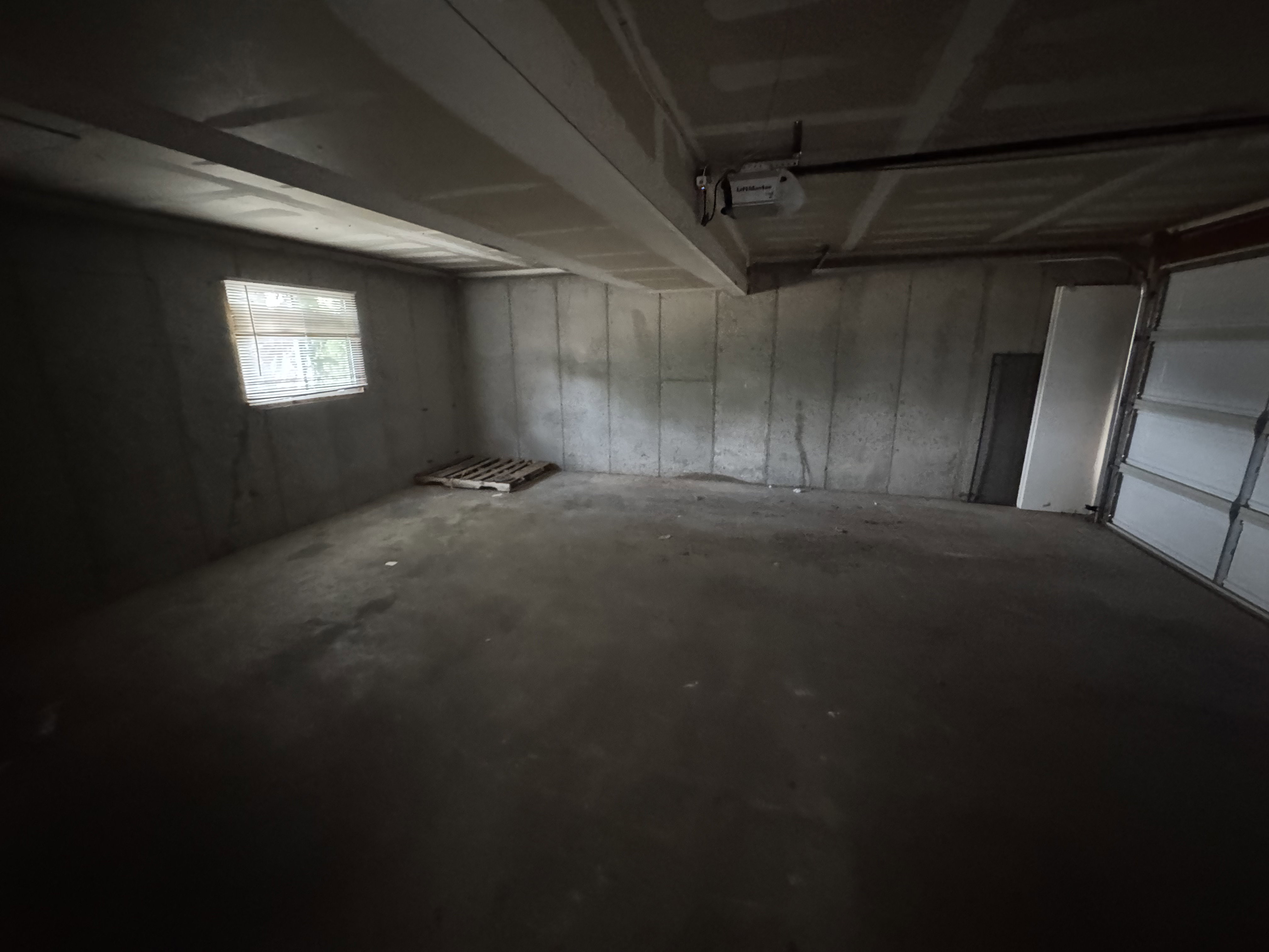 Concrete floor basement space with a single window, exposed ceiling joists, concrete block walls, and a visible garage door