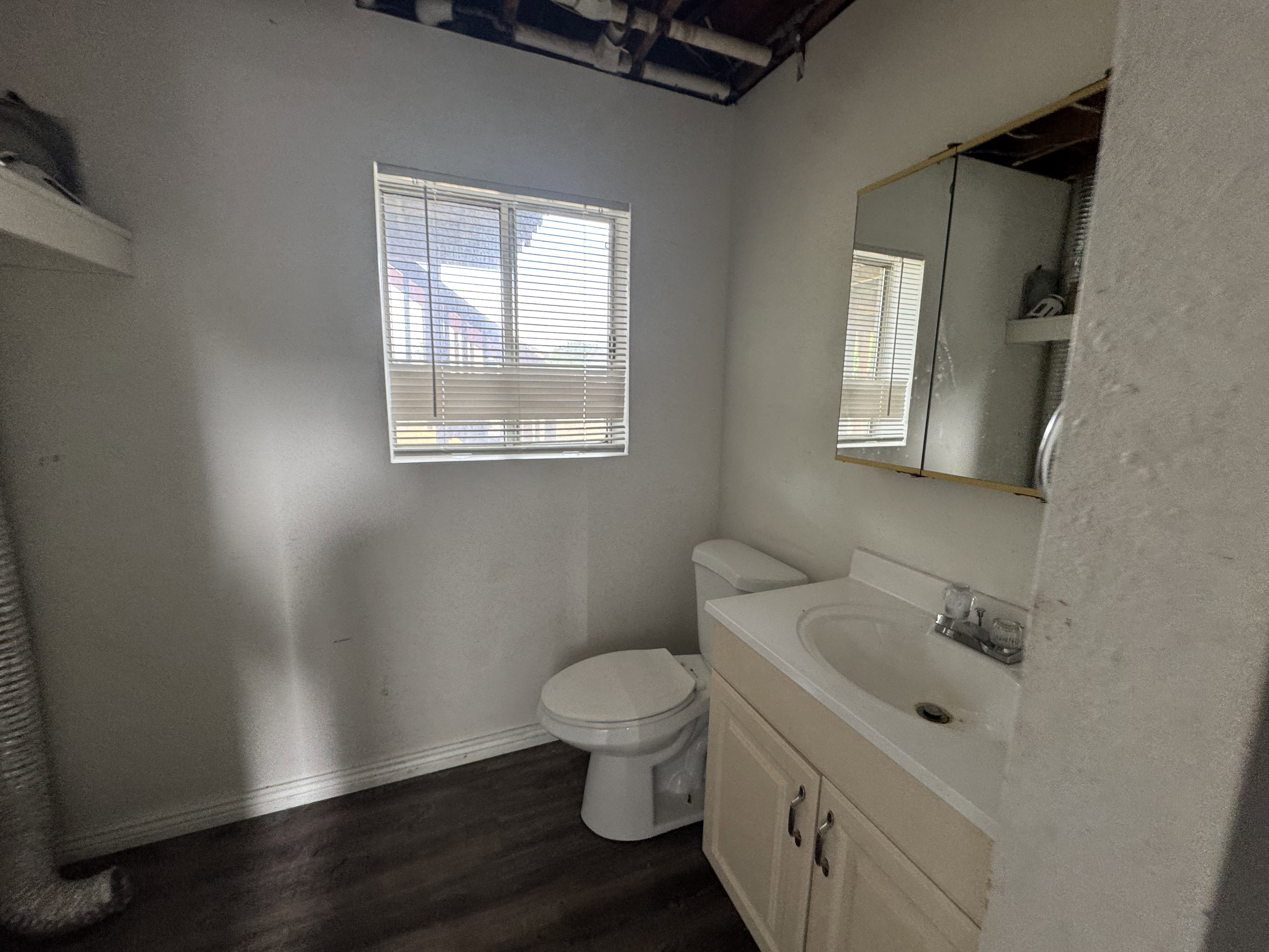 Wood-finish flooring bathroom featuring a single window, white toilet, vanity with integrated sink, and mirrored medicine cabinet