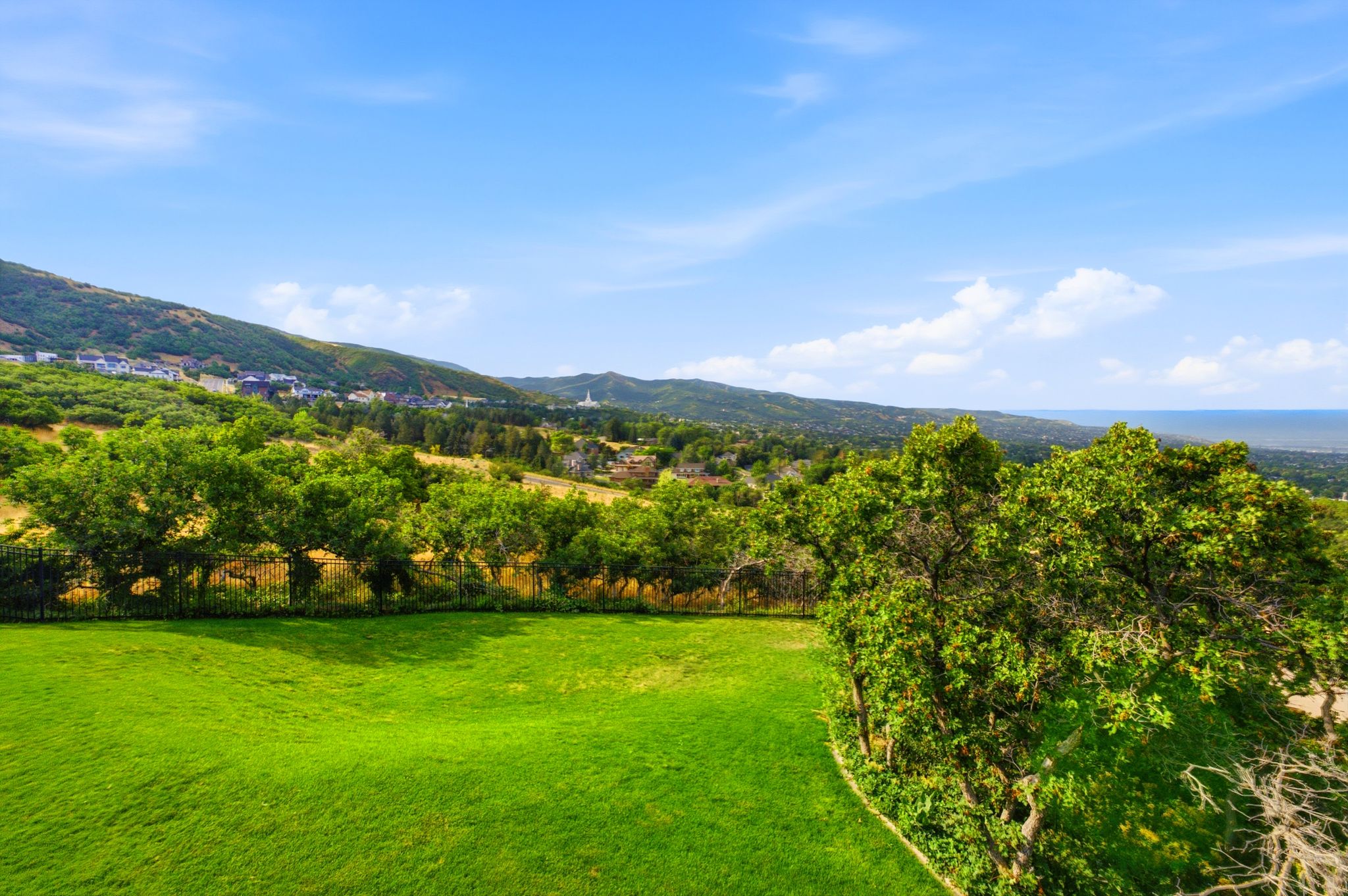 Expansive green lawn with mature trees and a black metal fence, complemented by distant hillside homes and a visible body of water