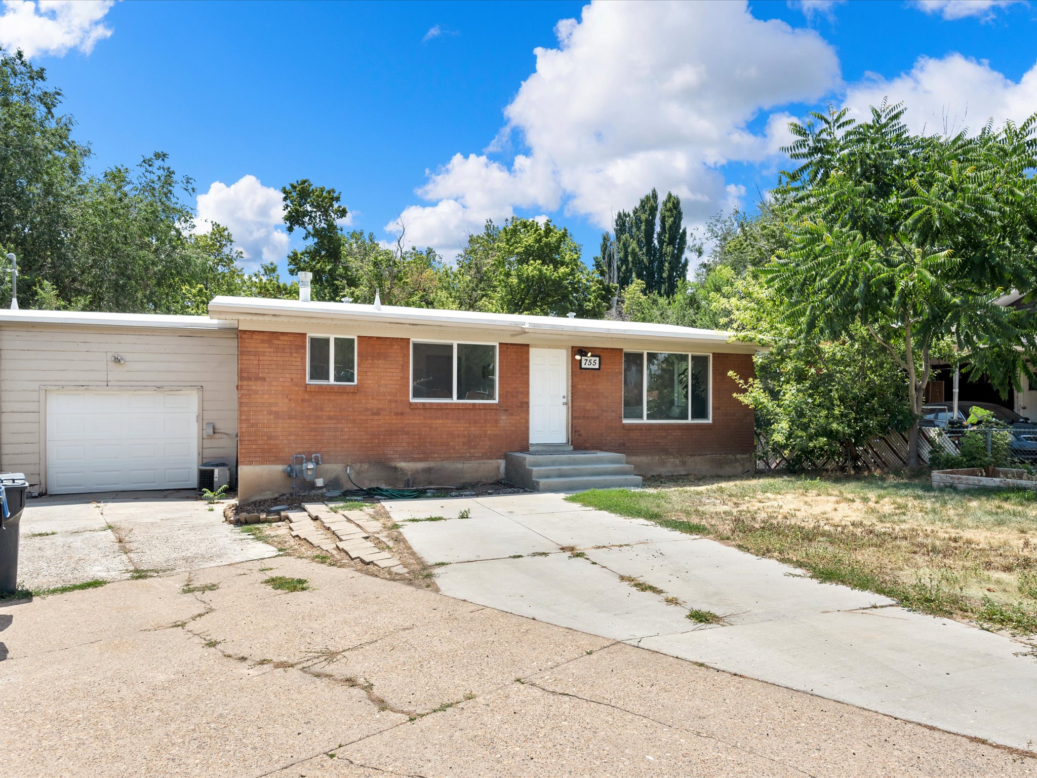 Brick exterior residence with an attached garage, a white entry door, multiple windows, a concrete driveway, and established trees