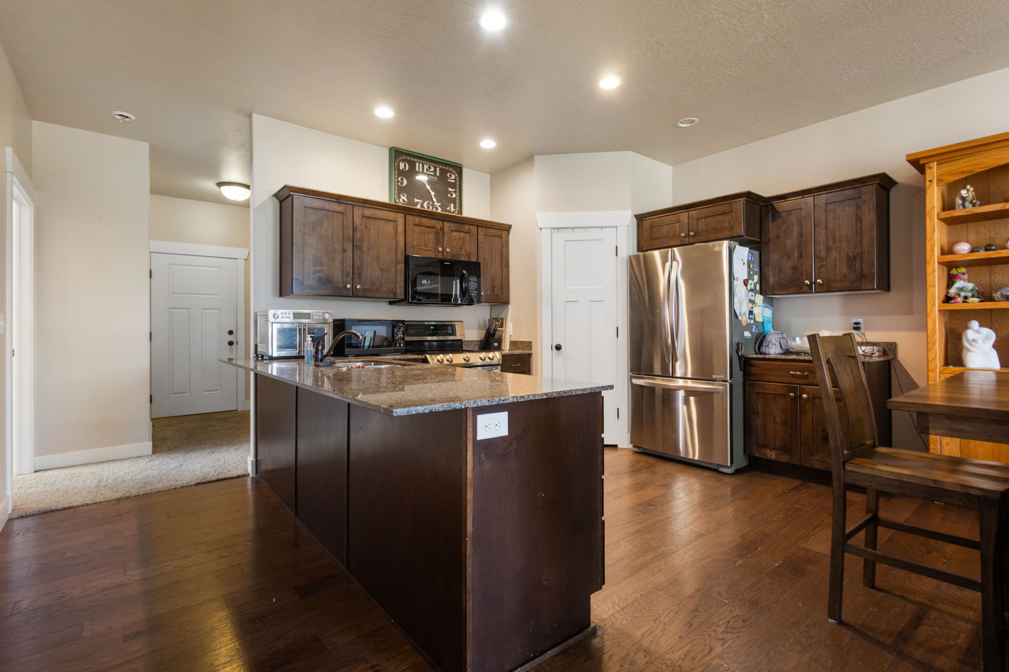 Kitchen featuring wood-finish flooring, dark wood cabinetry, stainless steel refrigerator, black appliances, and a granite-topped island with electrical outlets