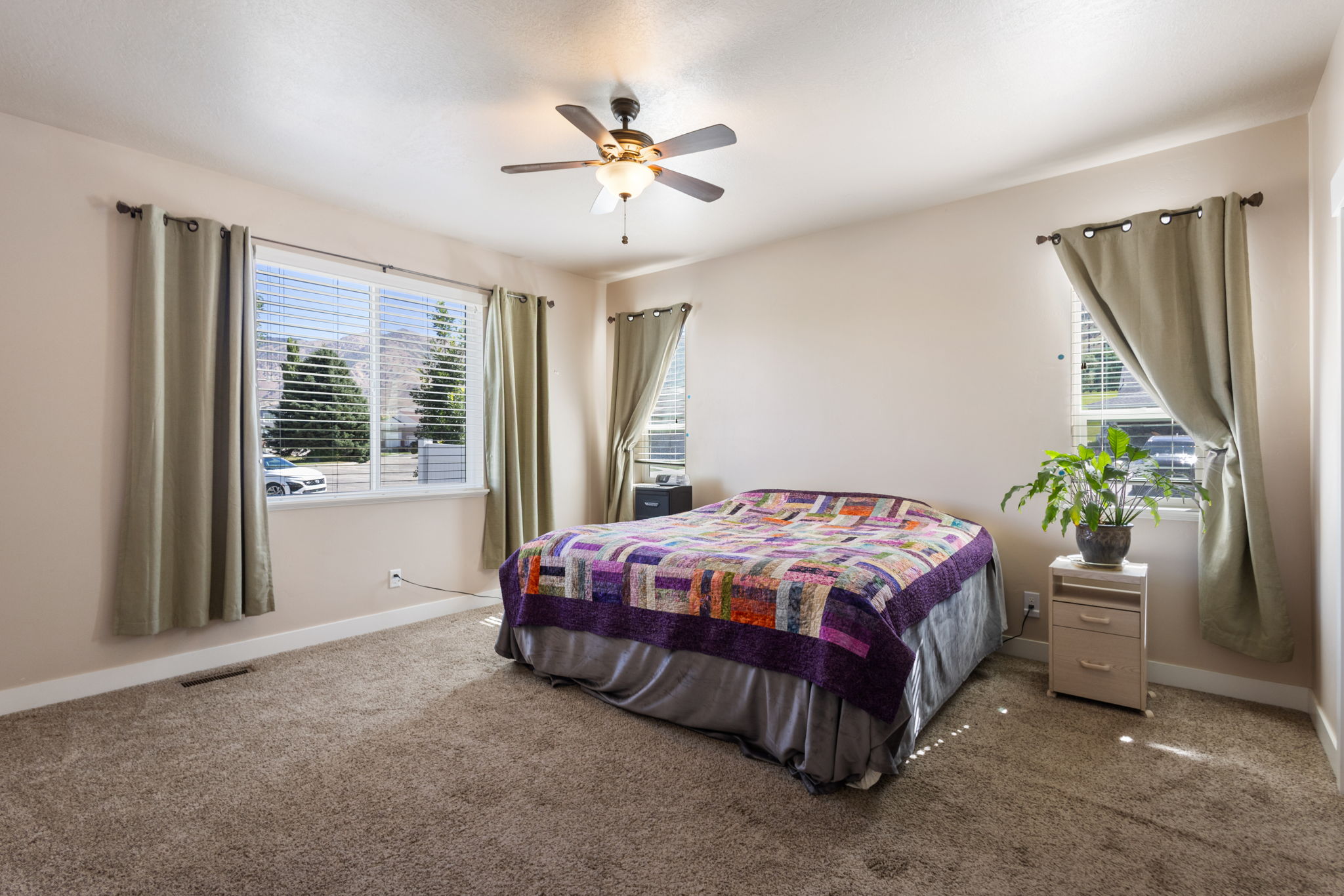Carpeted bedroom featuring two windows with horizontal blinds, a ceiling fan with integrated lighting, and neutral wall tones