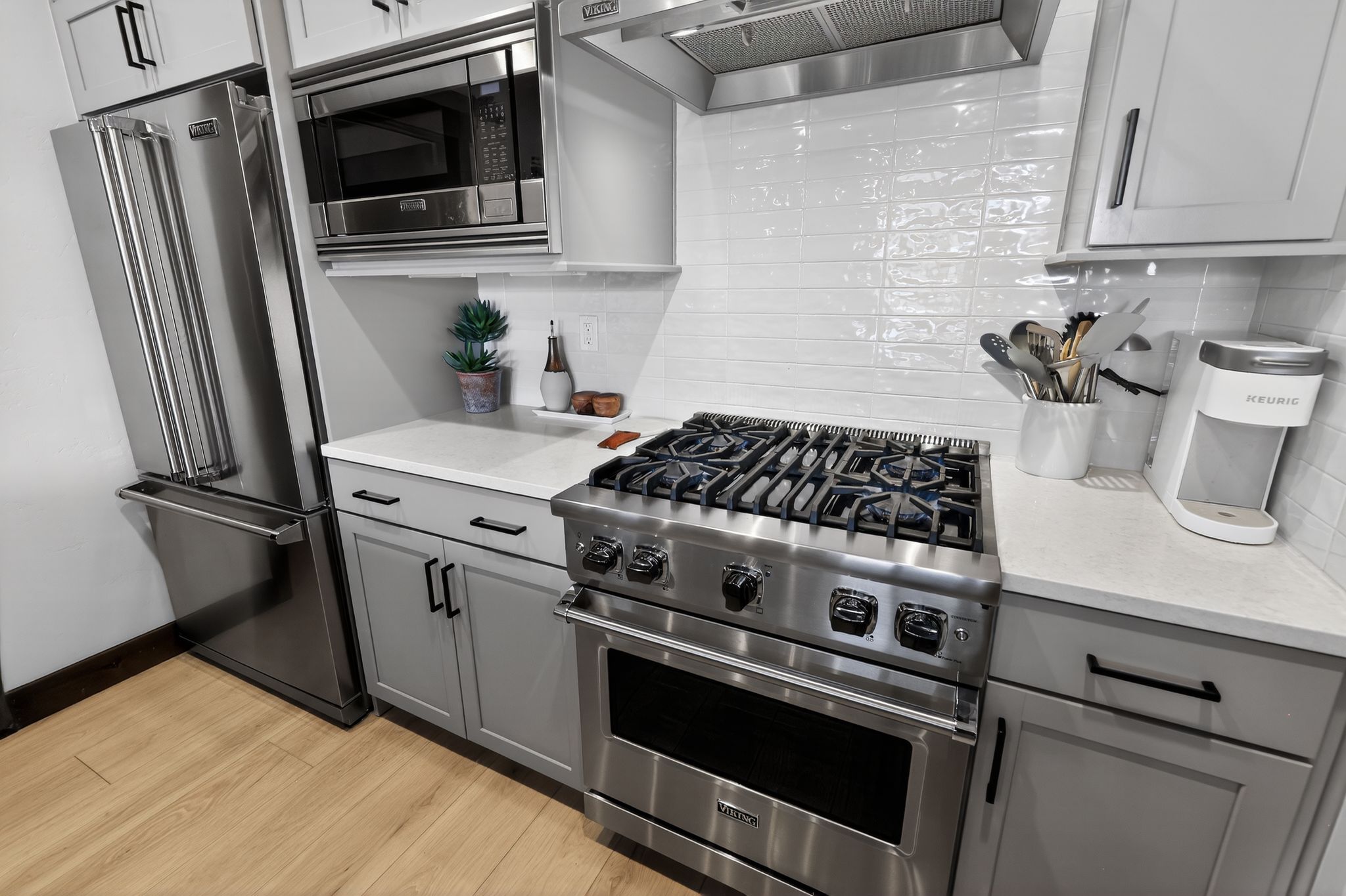 Kitchen featuring stainless steel appliances, light gray cabinetry with black hardware, white subway tile backsplash, light-toned countertops, and wood-finish flooring