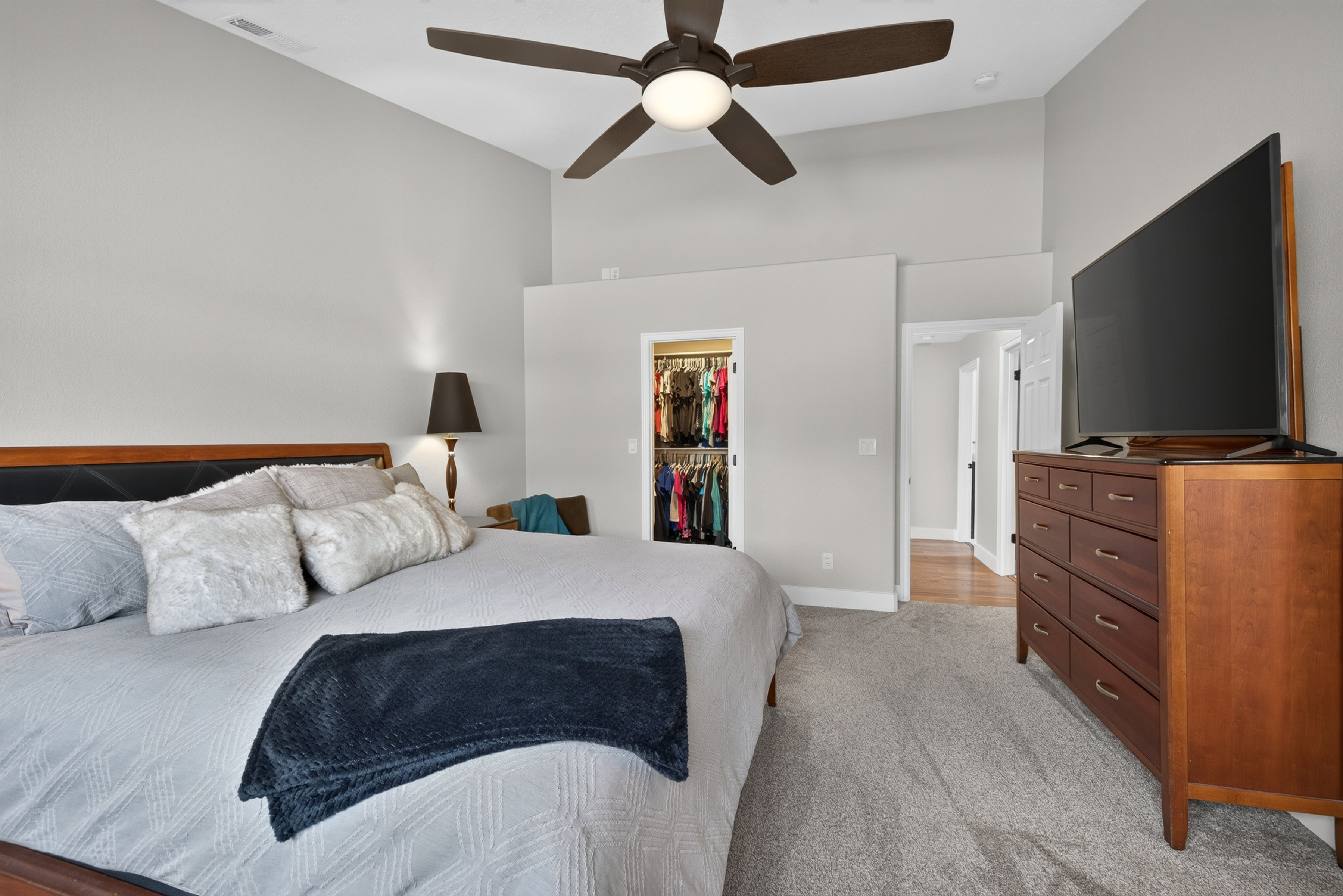 Vaulted ceiling bedroom featuring a ceiling fan with integrated lighting, soft gray wall tones, plush gray carpeting, and a walk-in closet with built-in shelving