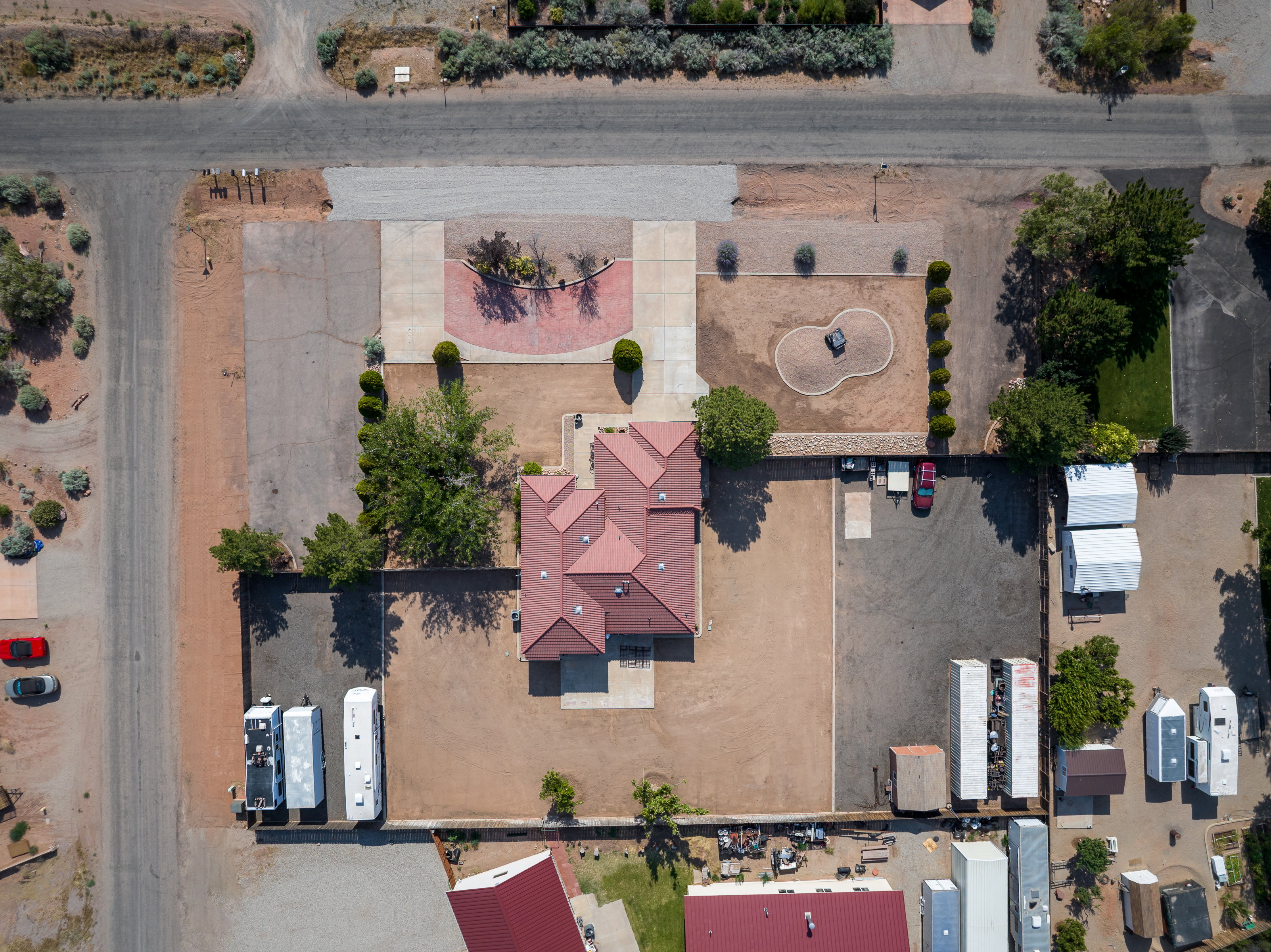 Expansive property featuring a red tile roof, concrete and gravel parking areas, mature trees, and a decorative brick-patterned circular drive