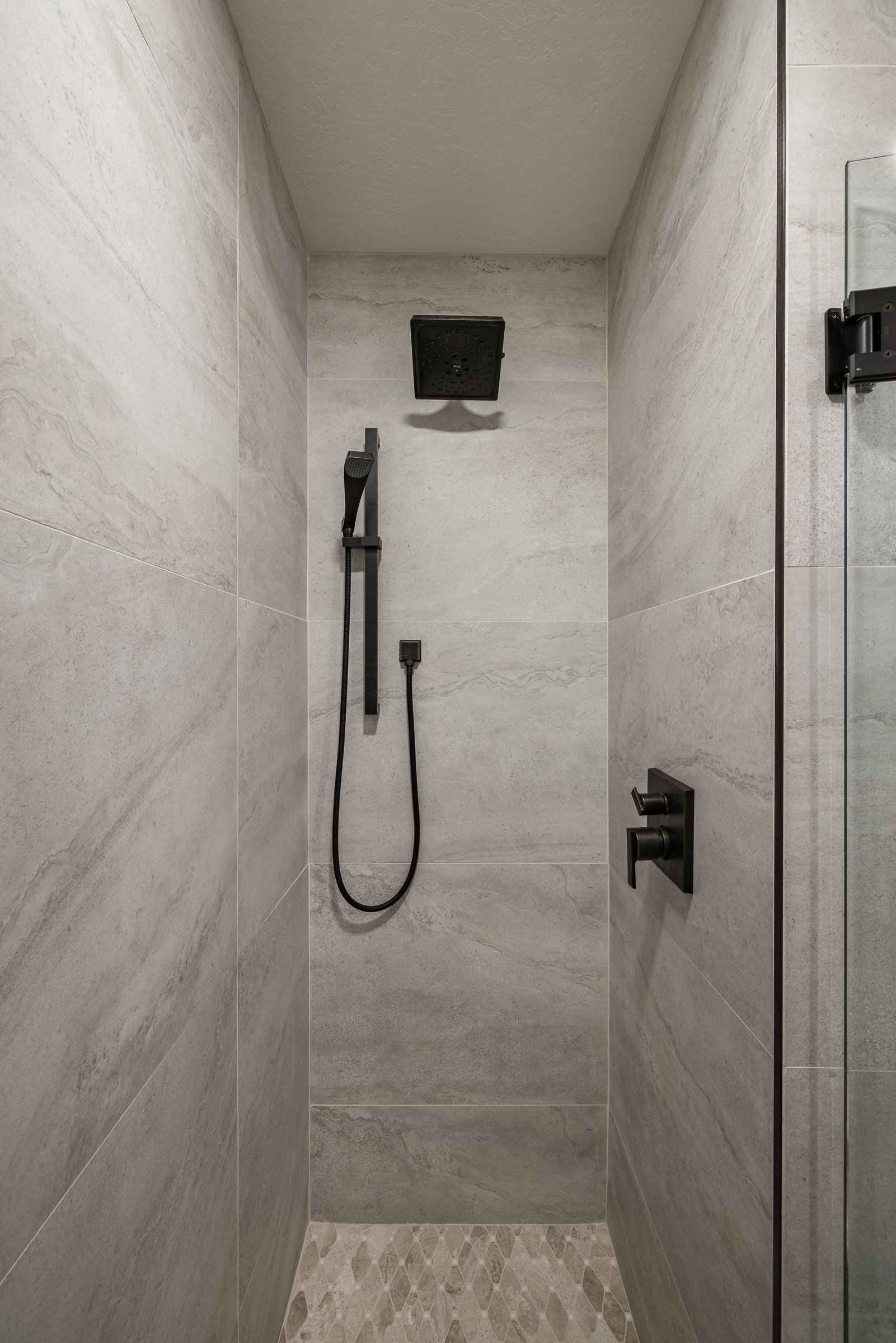 Shower enclosure featuring matte black fixtures, stone-look tile walls, and a decorative pebble-style tile floor