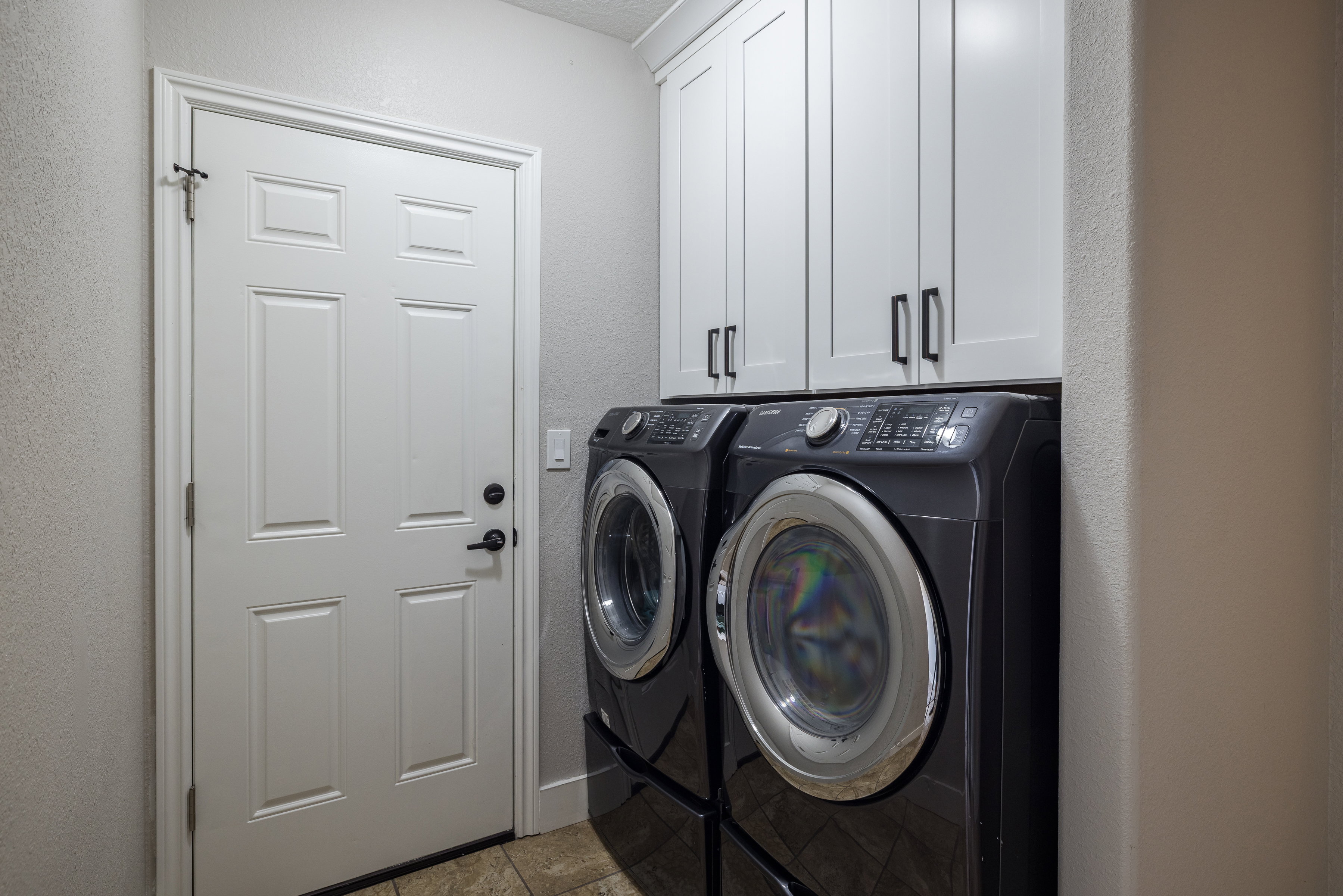 Laundry area featuring tile flooring, white shaker-style cabinetry with matte black hardware, and a white paneled door with matte black hardware