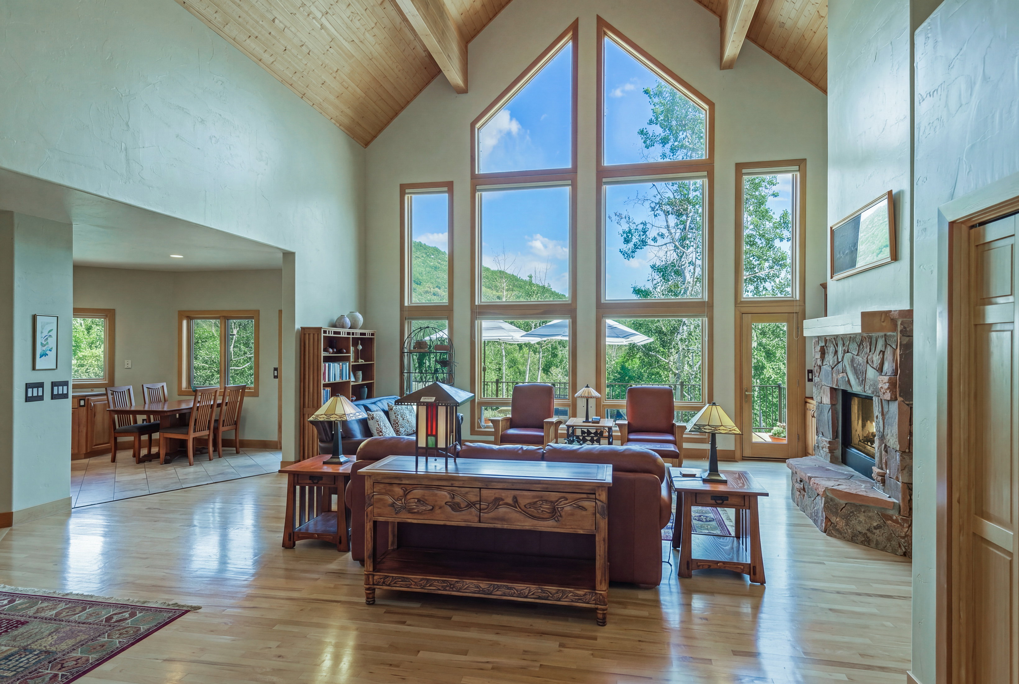 Vaulted ceilings with exposed wood beams define this spacious living area, featuring a stone-faced fireplace, light wood-finish flooring, and a dining area with tile flooring