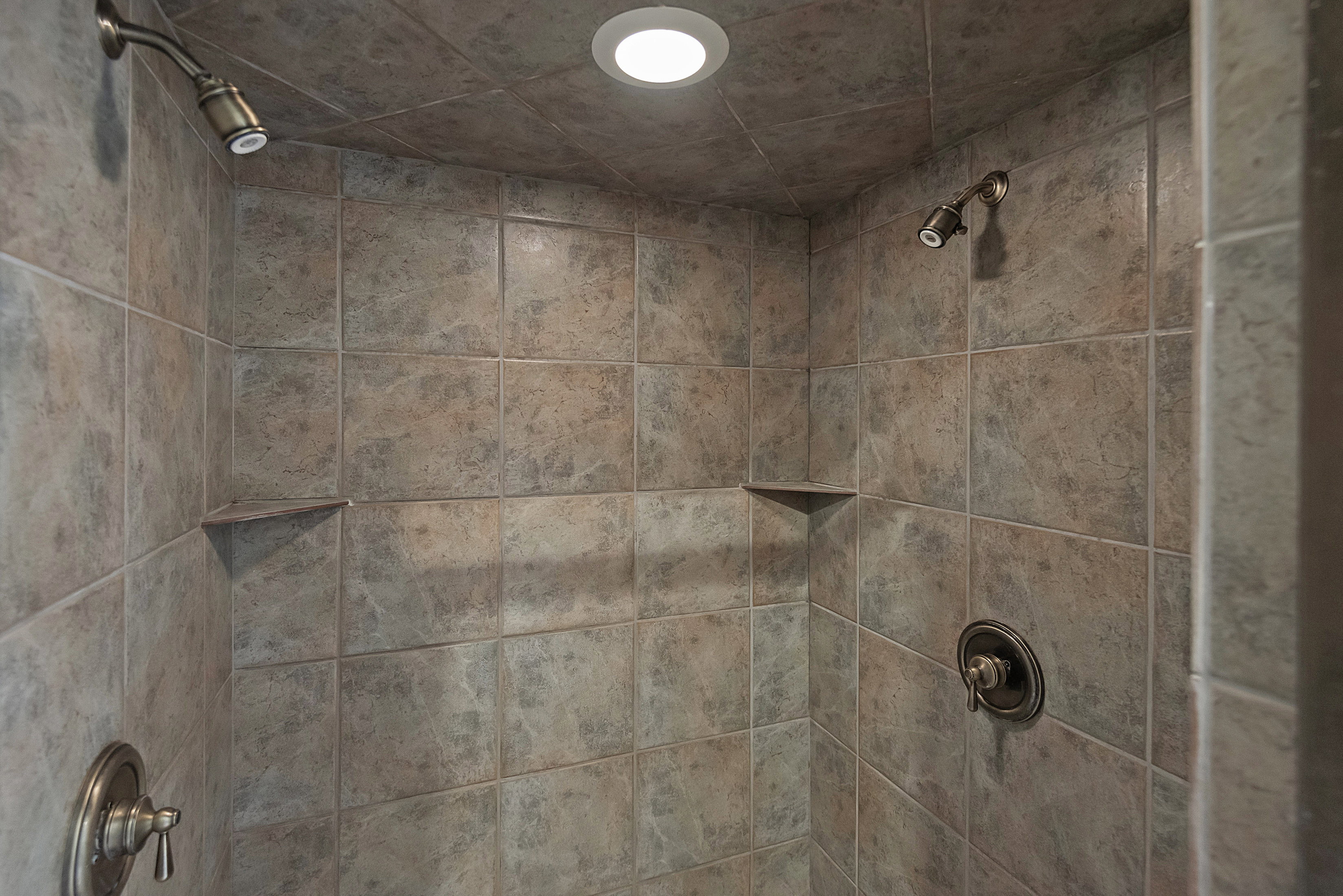 Tiled shower enclosure featuring dual shower heads with bronze-finish fixtures, recessed lighting, and integrated corner shelves