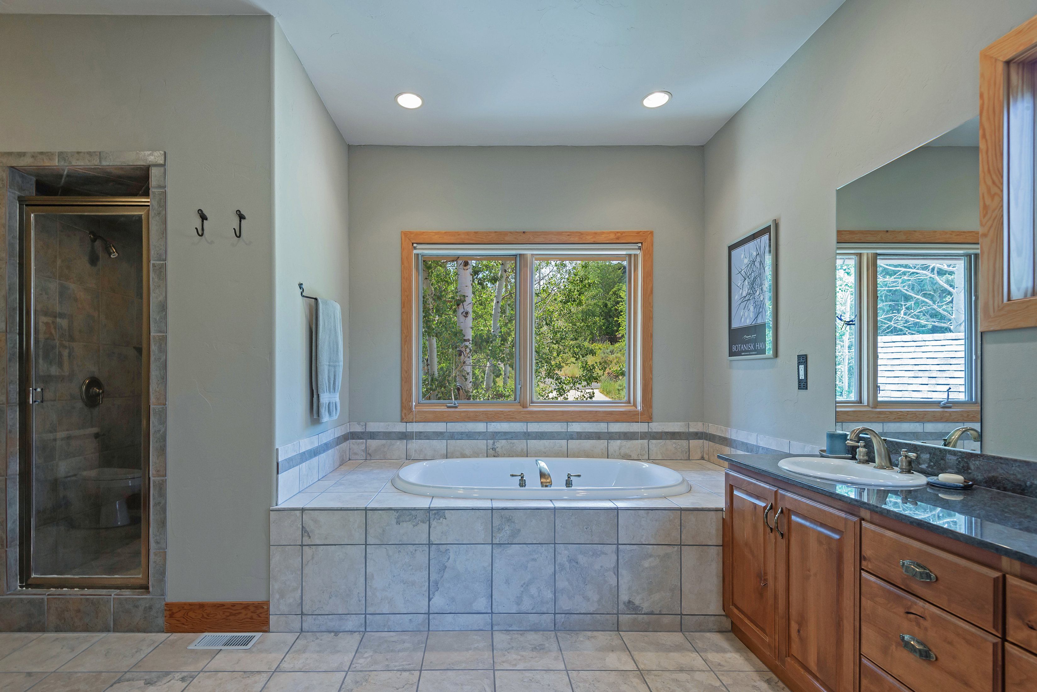 Bathroom featuring a large soaking tub with tile surround, a separate glass-enclosed shower with tile walls, a vanity with wood cabinetry and dark countertop, and a window with wood trim