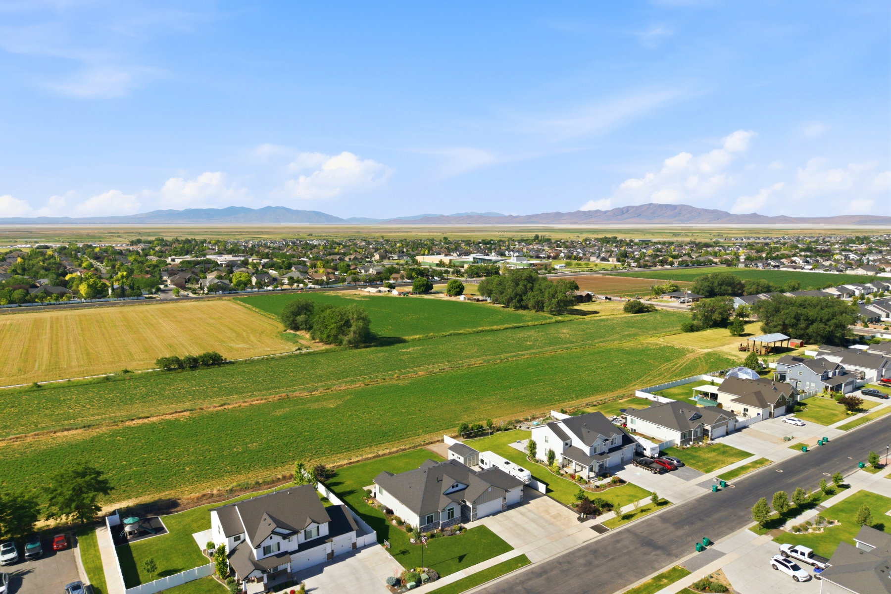 Aerial perspective showcasing a residential street with single-family homes, expansive green fields, and distant mountains under a clear sky