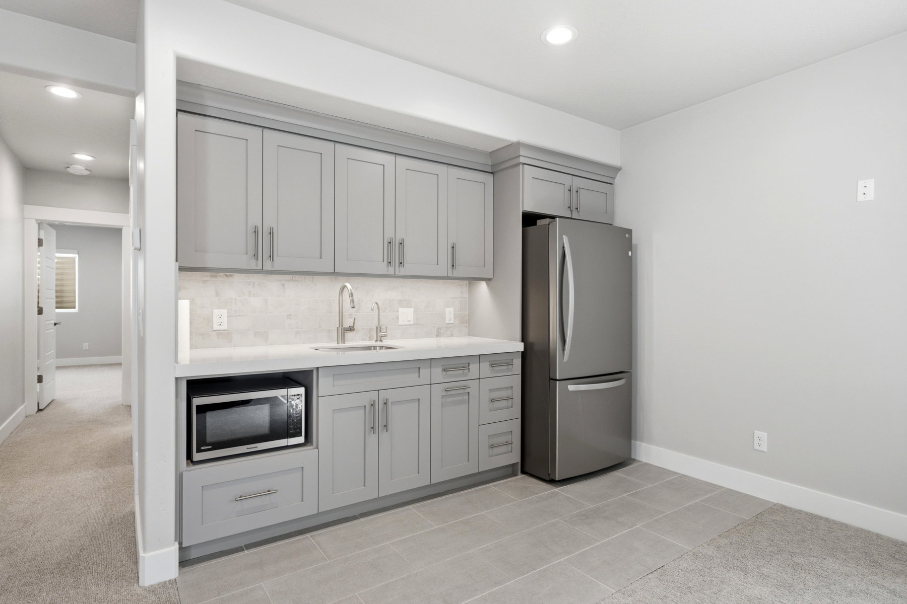 Lower level kitchenette featuring gray shaker-style cabinetry, a built-in microwave, stainless steel refrigerator, undermount sink, and light-toned tile backsplash