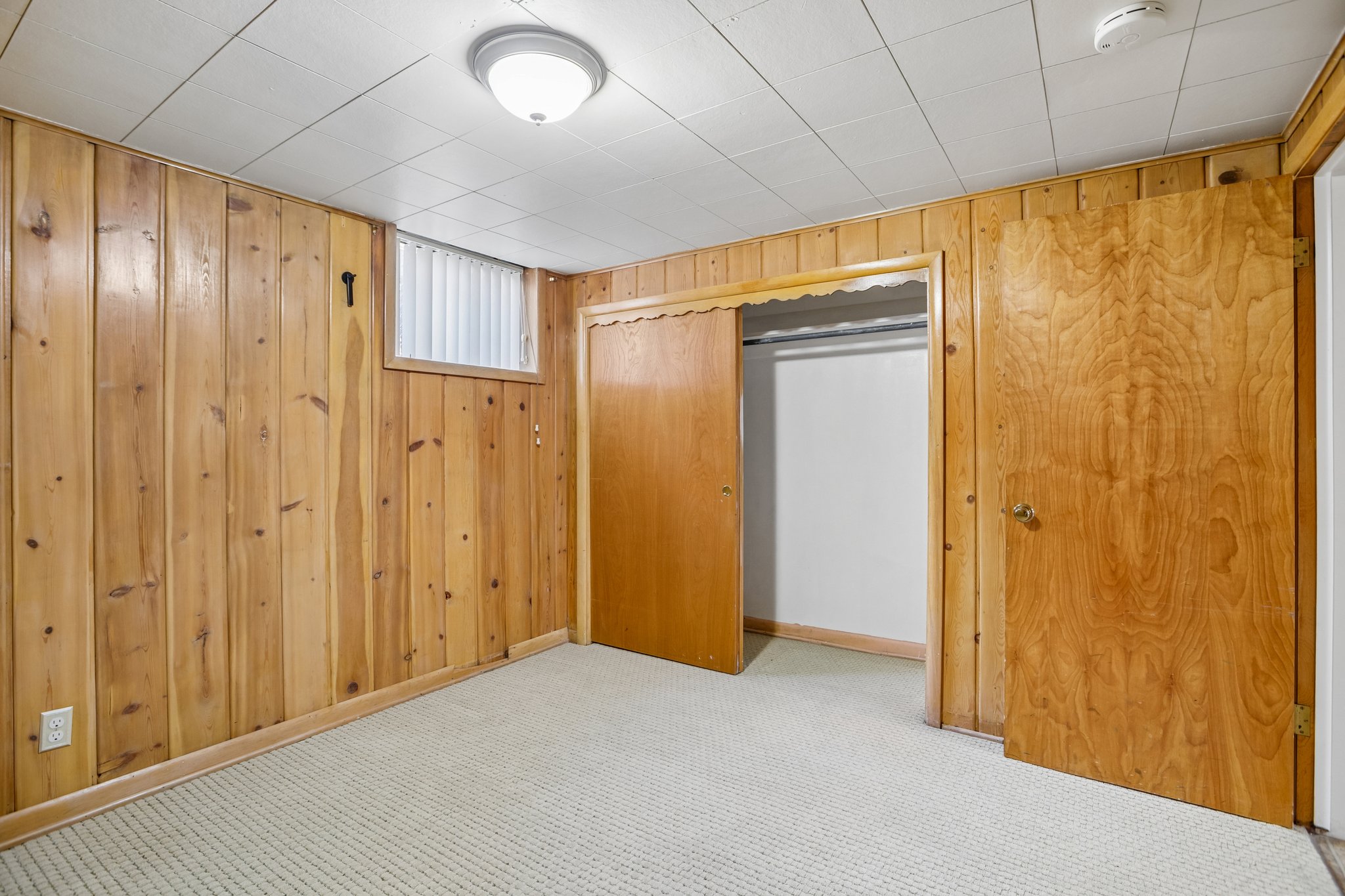 Room featuring wood paneling, light-colored carpet, a flush-mount ceiling light, a built-in closet with sliding wood doors, and a window with blinds