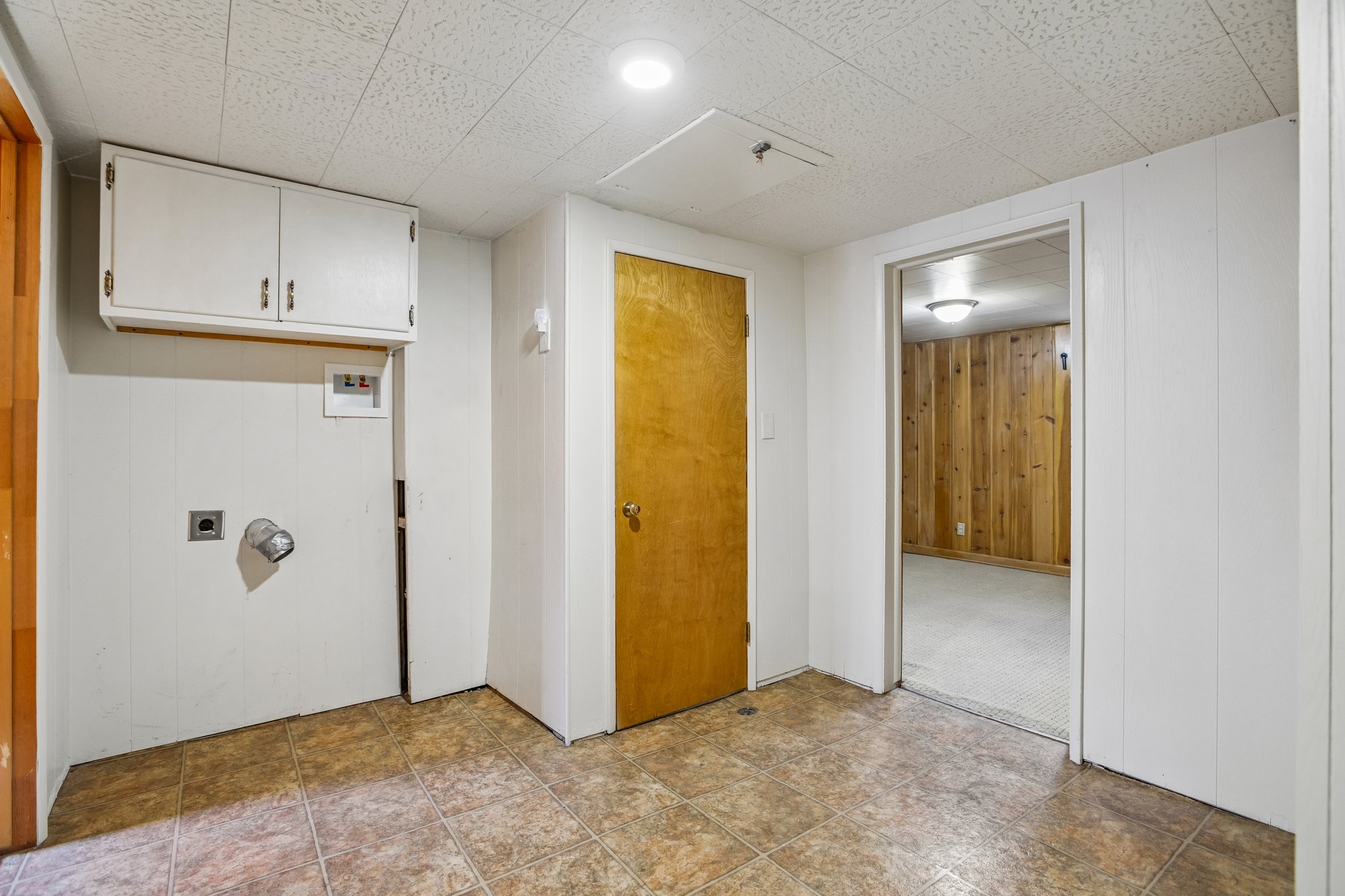 Laundry area featuring brown tile flooring, white panel walls, and upper cabinetry
